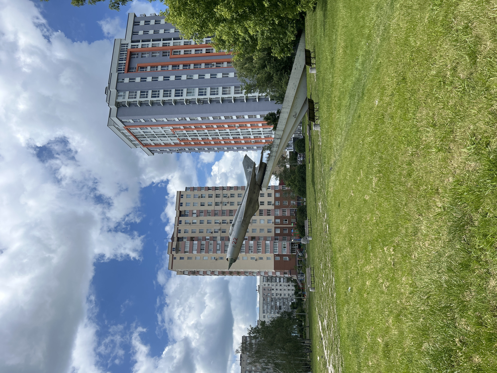 This image shows a memorial to a MiG-21 fighter jet, likely located in Bila Tserkva, Ukraine. The jet is mounted on a concrete stand in a grassy area. In the background, there are several tall residential buildings and trees. The sky is partly cloudy with a bright blue sky visible. The memorial appears to be in a public space, possibly a park or a residential courtyard, as there are benches in the foreground. The jet has the number "22" painted on its nose. The overall impression is a somewhat weathered but still striking monument in an urban setting.