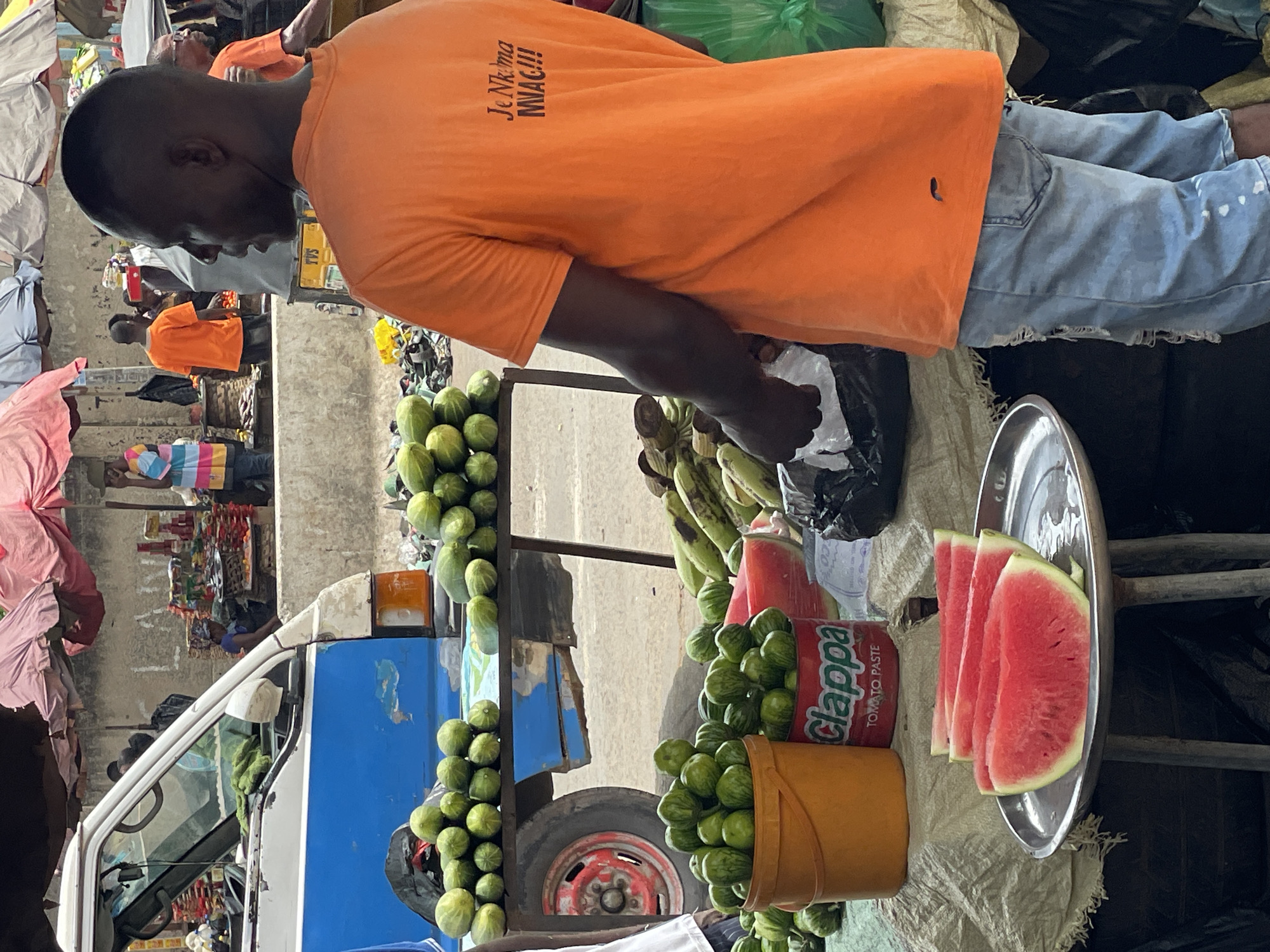 An eye-level medium shot depicts a street vendor with dark skin, an orange t-shirt, and jeans, reaching into a plastic bag. His back is to the camera, and the text "Je Nk'ma MWAC!!!" is printed on his shirt. In front of him is a display of produce and processed goods. A silver tray holds four sliced watermelons. A yellow bucket is overflowing with small, green gourds, and a large Clappa Tomato Paste can is visible between them. Additional produce, including bananas and more of the same type of gourds, is arranged nearby. A blue and white pickup truck is partially visible behind the vendor's display, with more gourds attached to it. The scene appears to be a marketplace, potentially located in the city of Onitsha, Nigeria. Other people are present in the background, suggesting a bustling street scene.