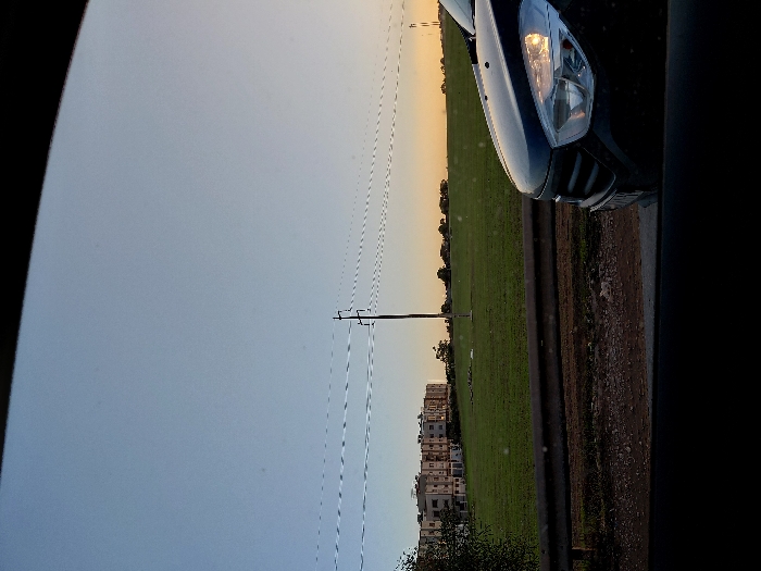 A car's headlight shines in this outdoor shot. A green field extends along the horizon, punctuated by buildings and power lines. The sky above is overcast, transitioning from a lighter hue near the horizon to a darker shade overhead. Part of the car's body is visible, seemingly taken from inside the vehicle. The overall image evokes a sense of travel and observation.