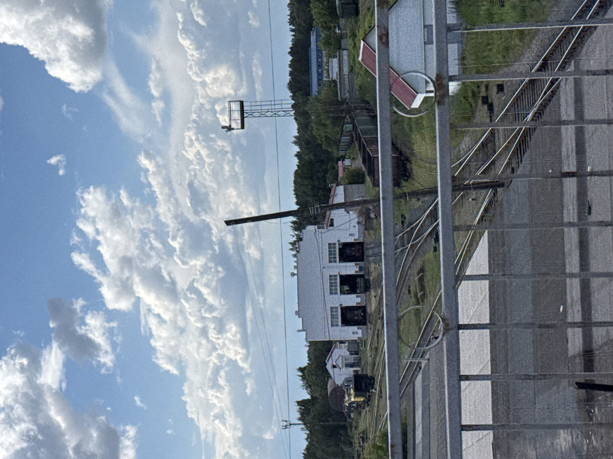 The image shows a railway yard in Chaikovsky, Russia. In the foreground, there is a grey metal fence. Behind the fence, numerous railway tracks are visible, along with some grassy areas and low vegetation. In the middle ground, several railway buildings are present, including a large white building with large open doorways where trains appear to be housed. To the left of the large white building, there are smaller buildings and a yellow and black train engine. Further into the background, a long line of dark-colored train cars is visible on a track. Beyond the railway yard, a dense line of green trees forms the horizon. Above the trees, a tall metal tower with a platform at the top stands against a bright blue sky filled with fluffy white clouds. The overall scene depicts an industrial area related to railway operations under a partly cloudy sky.