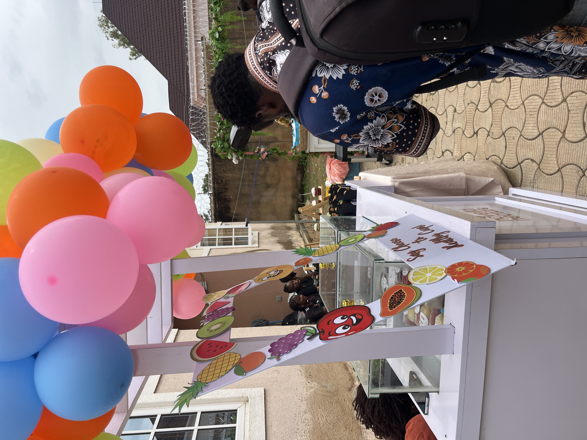 In the city of Bar Jirgi Summa, Nigeria, a mobile juice stand is set up outdoors. The stand is decorated with colorful balloons in shades of pink, blue, orange, and yellow, as well as cutouts of various fruits like pineapples, watermelons, grapes, apples, and papayas. A sign on the stand reads "Say Cheese to Healthy Cups!".
There are at least three people visible. Two women are seated behind the counter of the stand, appearing to be engaged in conversation. In the foreground, a person wearing a blue shirt with a floral pattern and a backpack is leaning over the counter, looking at the items displayed. The background shows a building with a dark brown roof and windows, and a fence with barbed wire. The ground is paved with interlocking concrete blocks. The overall scene suggests a vibrant and possibly festive atmosphere.