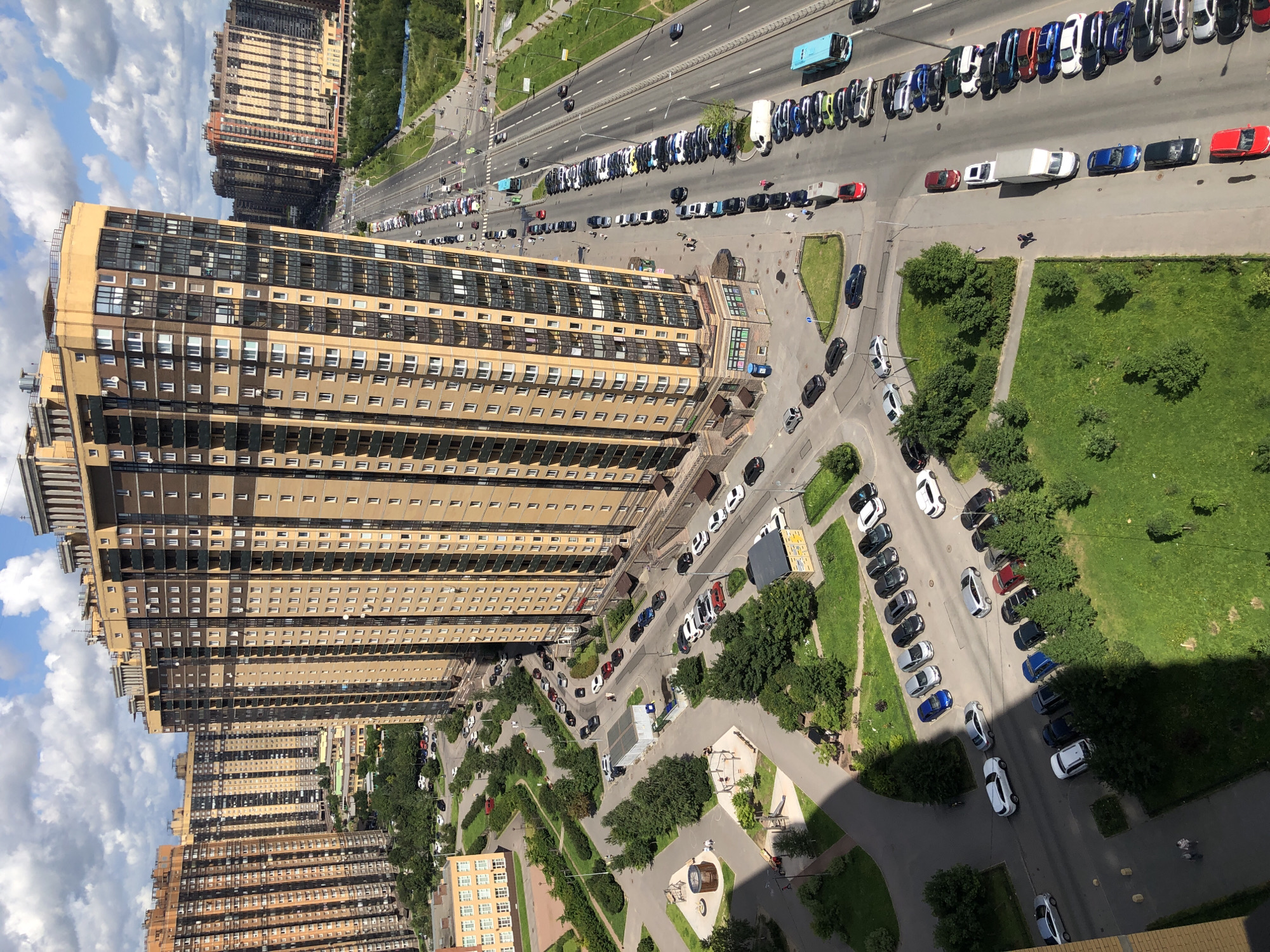 An aerial view captures a dense urban landscape dominated by towering residential buildings. In the foreground, a light brown and yellow high-rise apartment building features numerous windows and balconies. Adjacent to it, another similarly tall building with a darker facade is visible. A busy multi-lane road runs alongside these buildings, lined with parked cars of various colors and makes, including sedans, SUVs, and a blue bus. The road is divided by white lines and has traffic signals at intersections. Green spaces, including trees and manicured lawns, are interspersed throughout the urban environment. In the lower left quadrant, a playground area with swings and climbing structures is present, with two individuals walking nearby. The sky above is partly cloudy, with patches of blue visible.