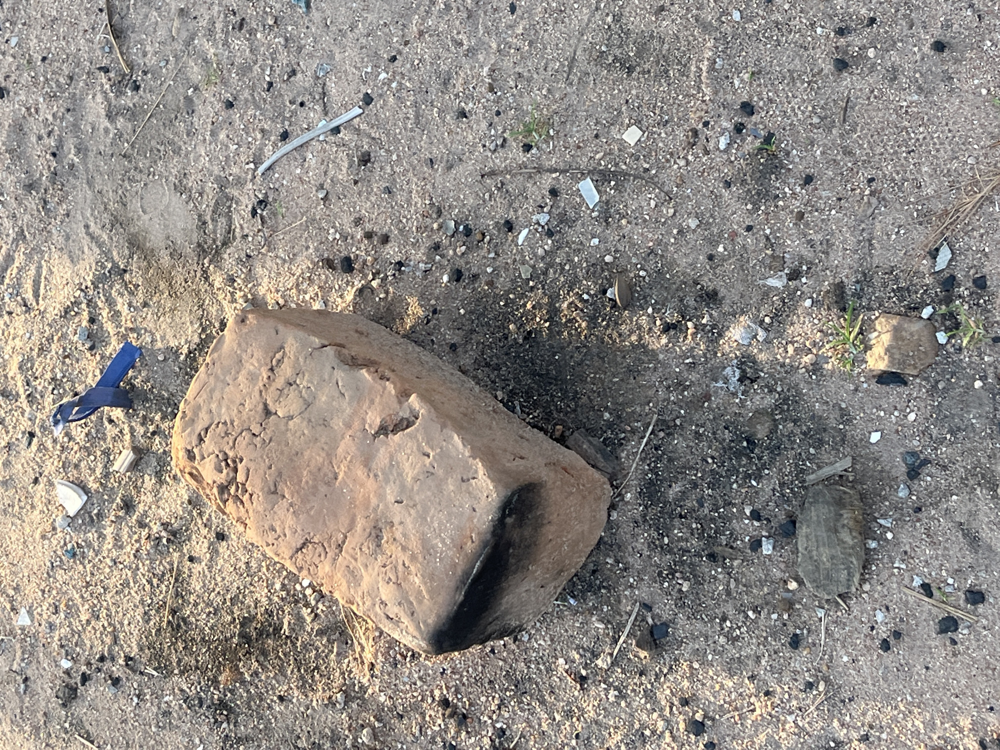 The image shows a close-up, overhead view of a brick lying on sandy ground littered with small pebbles, debris, and dried grass. The brick, which appears weathered and possibly burnt on one corner, is the most prominent object. To the left of the brick, a fragment of blue ribbon or plastic is visible. The ground is a mix of light brown sand and darker particles, possibly charcoal or small stones. In the background, more small rocks and sparse vegetation can be seen. The lighting suggests it is daytime, possibly late afternoon, given the soft shadows cast by the brick. The scene appears to be outdoors in a dry, uncultivated area. No people are visible, nor is there any text.