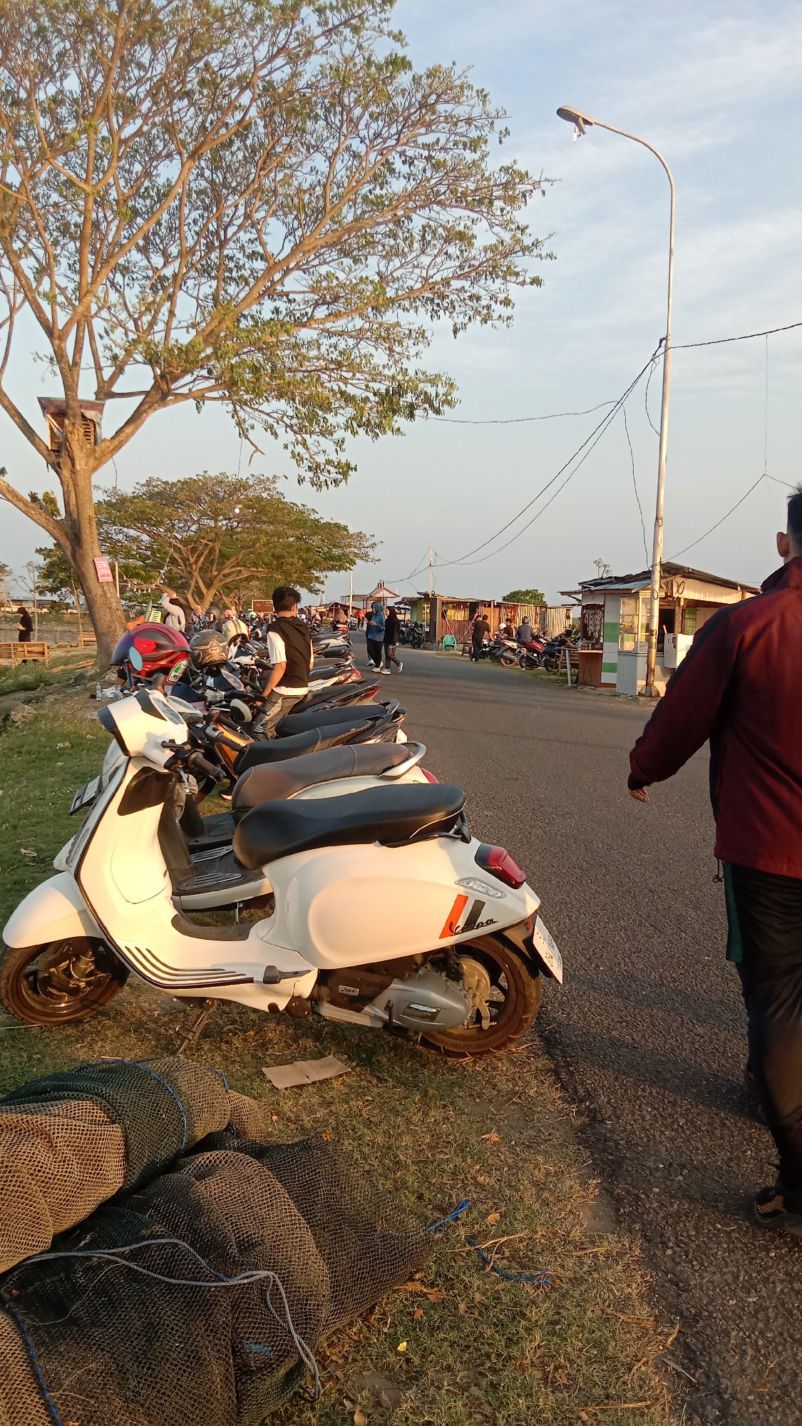 The image shows a roadside scene near a waterfront in Lhokseumawe, Indonesia. A row of parked motor scooters, mostly Vespa-style, lines the edge of a paved road. To the right, a person in a maroon jacket walks away from the camera. In the background, people stroll along the road and gather near small structures that appear to be shops or stalls. A tall utility pole stands near the shops. A tree stands on the left side, with a birdhouse-like structure in its branches. In the lower left corner, fishing nets are piled on the grass.
