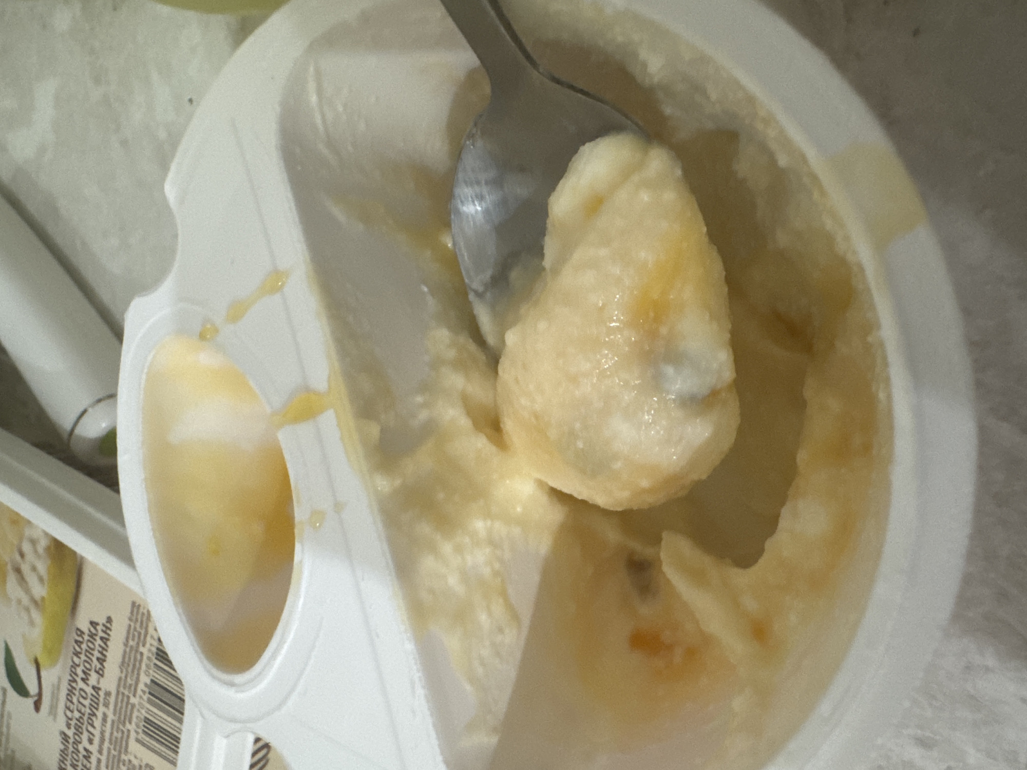 The image shows a close-up of a white plastic container with a yellow, creamy substance that appears to be some kind of food product. A metal spoon is partially submerged in the substance, scooping up a portion of it. The container has a divided section at the top, and the food product fills the lower section. There is some of the yellow substance also visible on the rim of the container, suggesting it has been eaten from. Part of a label with Russian text and a barcode is visible in the upper left corner of the image. The background is out of focus but appears to be a textured surface, possibly a table. The lighting is somewhat dim.
Without any additional information or context, it is impossible to determine the location of this image within the city of Saratov, Russia. The image itself does not contain any visual cues that would indicate a specific geographic location.