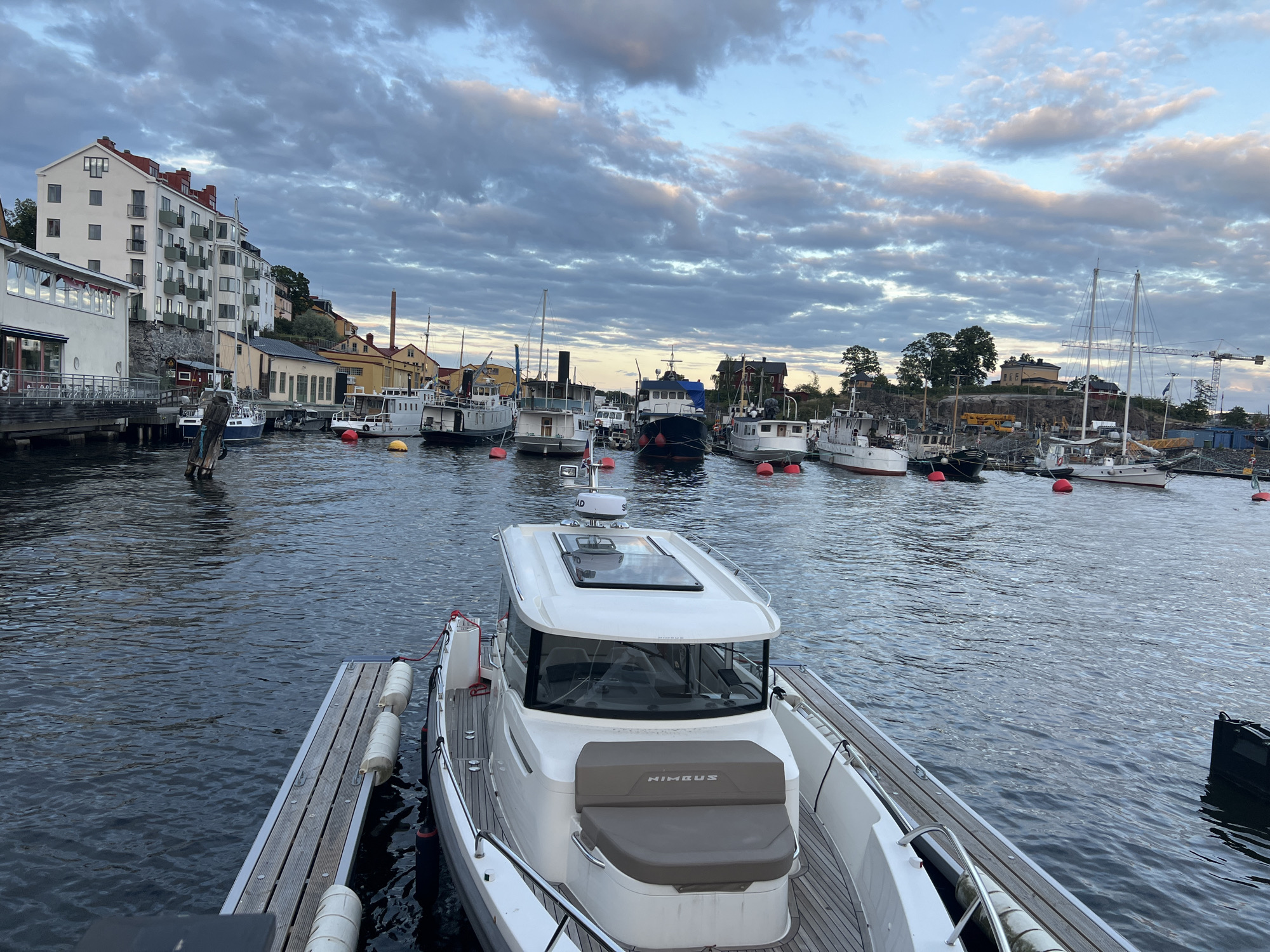 A fleet of various boats, ranging from small pleasure craft to larger vessels, are docked in a tranquil harbor against the backdrop of Stockholm's urban landscape. In the foreground, a modern white motor yacht, identified by the "NIMBUS" branding on its seating, is moored to a wooden pier. The boat features a sleek cabin with large windows and a skylight. Beyond the yacht, a collection of other boats are lined up, some with masts and sails, others appearing to be working vessels or older motorboats.
The cityscape on the left side of the image is characterized by multi-story buildings with white facades and red roofs, typical of Scandinavian architecture. A long, low-rise building with extensive windows and outdoor seating suggests a restaurant or cafe. The waterfront is lined with old industrial buildings, a tall brick chimney, and some greenery. The sky above is dramatic, with heavy clouds in shades of gray and blue, tinged with the soft light of either dawn or dusk, creating a moody atmosphere.
The water in the harbor is a deep, calm blue, reflecting the overcast sky and the silhouettes of the boats. Several red buoys are visible in the water, marking the boundaries of the harbor or navigational channels. The scene evokes a sense of peaceful coexistence between urban development and maritime life in Stockholm.