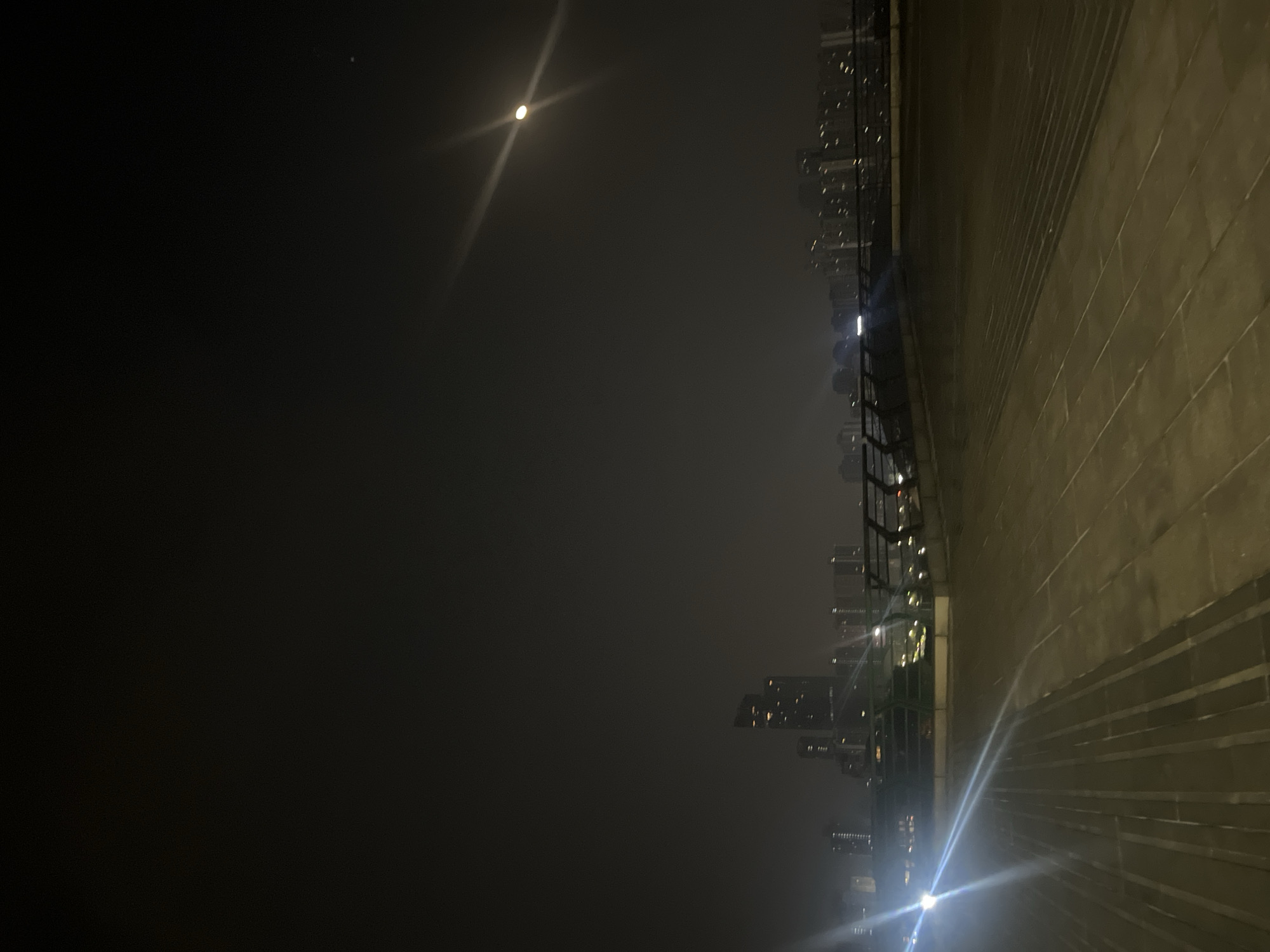 The image captures a nocturnal urban scene in Jiulongpo District, China. The foreground consists of a broad, multi-level paved area, illuminated by a strong artificial light source originating from the lower left, producing prominent lens flares across the pavement. The ground surface appears to be constructed from large, light-colored rectangular tiles or slabs. In the midground, a dark metal barrier or railing system is present, suggesting an elevated walkway or bridge. The background reveals a cityscape with numerous buildings, including various high-rise structures and residential blocks, many displaying illuminated windows, set against a dark, hazy sky. A distinct bright circular object, consistent with the moon or a powerful distant light, is visible in the upper right, also generating a prominent lens flare. No people, active subjects, or specific interactions are depicted.