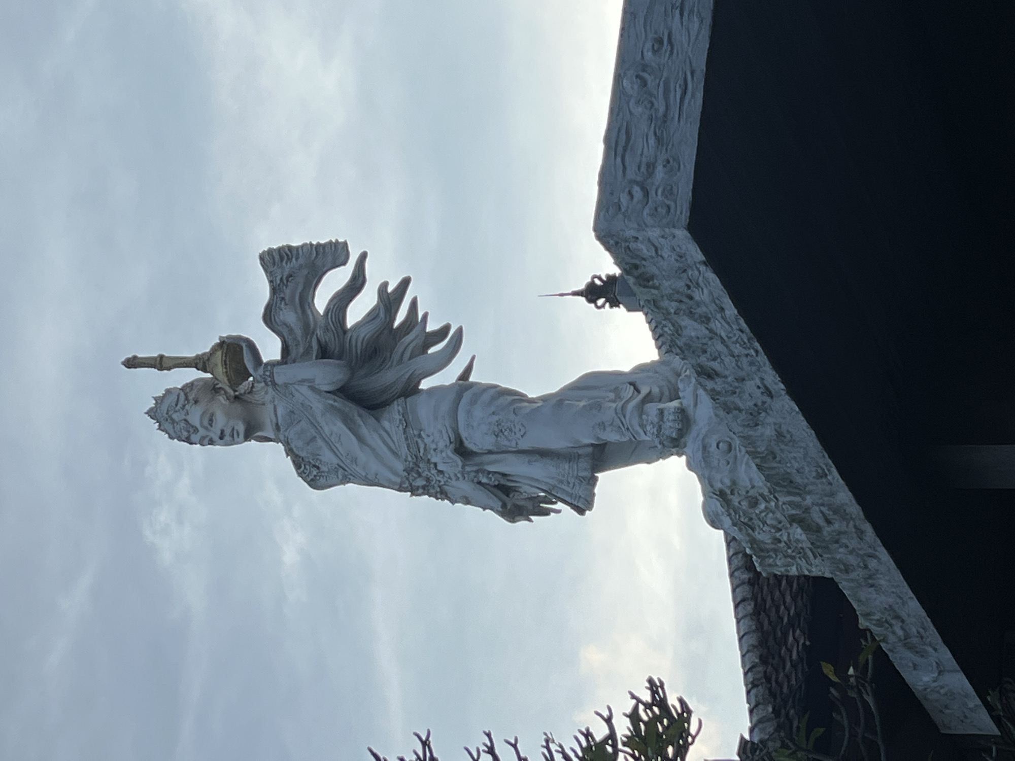 A low-angle shot shows a white statue of a female figure with a headdress and ornate wings, holding a cylindrical object above her head. The statue is mounted on a decorated white structure that appears to be part of a building, set against a cloudy sky. The building has a dark, contrasting element. The scene is in Sukawati, Indonesia.