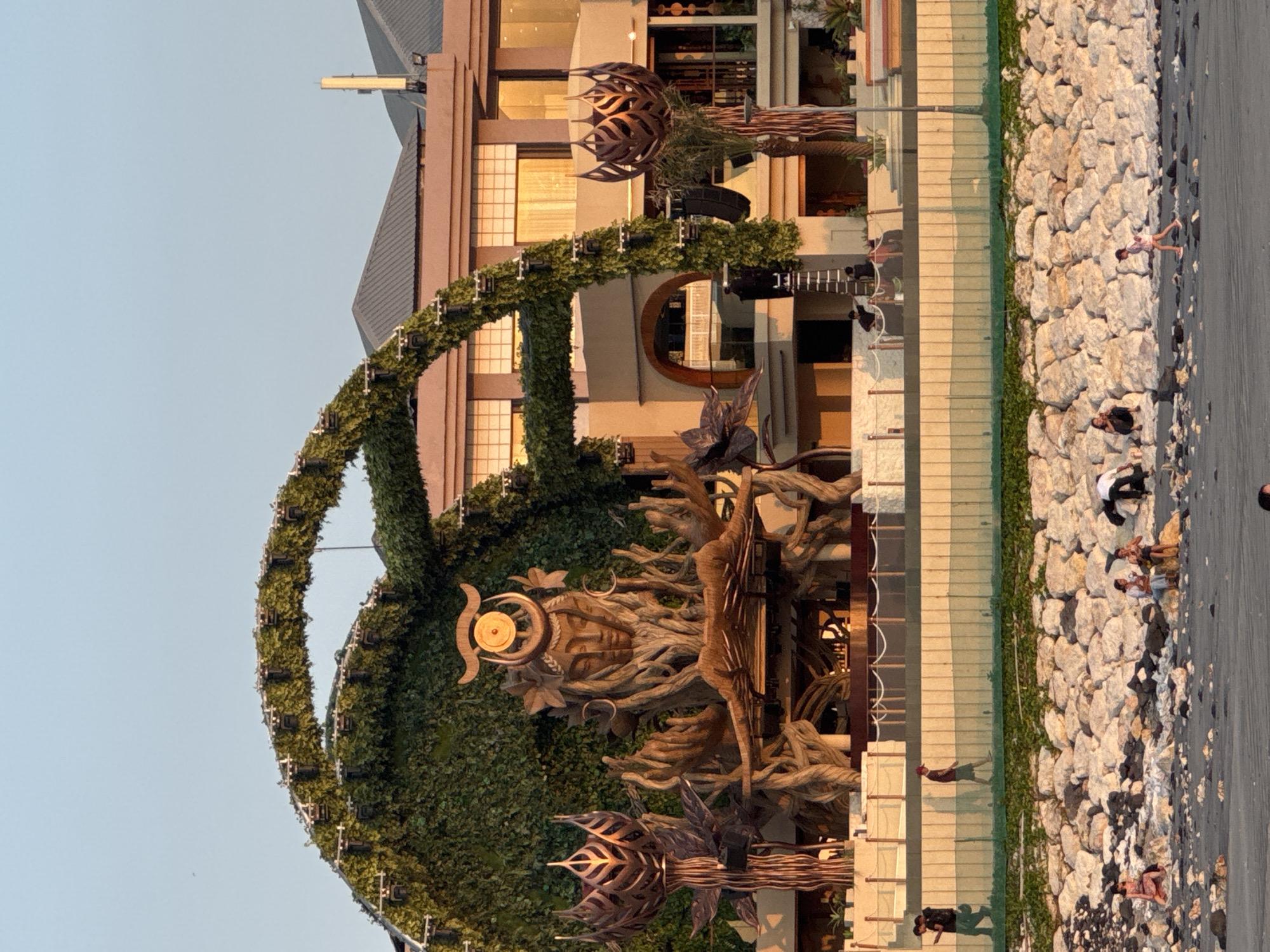 The image shows an outdoor stage setup in Kuta, Indonesia. The stage features a large, intricate sculpture resembling a face emerging from tree-like structures, adorned with leaves and branches. An arched canopy covered in greenery spans over the stage, with what appear to be lighting fixtures attached. Behind the stage, a modern building with multiple stories and large windows is visible, reflecting the warm light of what seems to be either sunrise or sunset. The foreground shows a beach area with a rocky embankment. Several people are scattered across the beach, some sitting, some walking, and a few are near the water's edge. The scene appears to be set for an event, possibly a music festival, given the stage design and audio equipment visible.