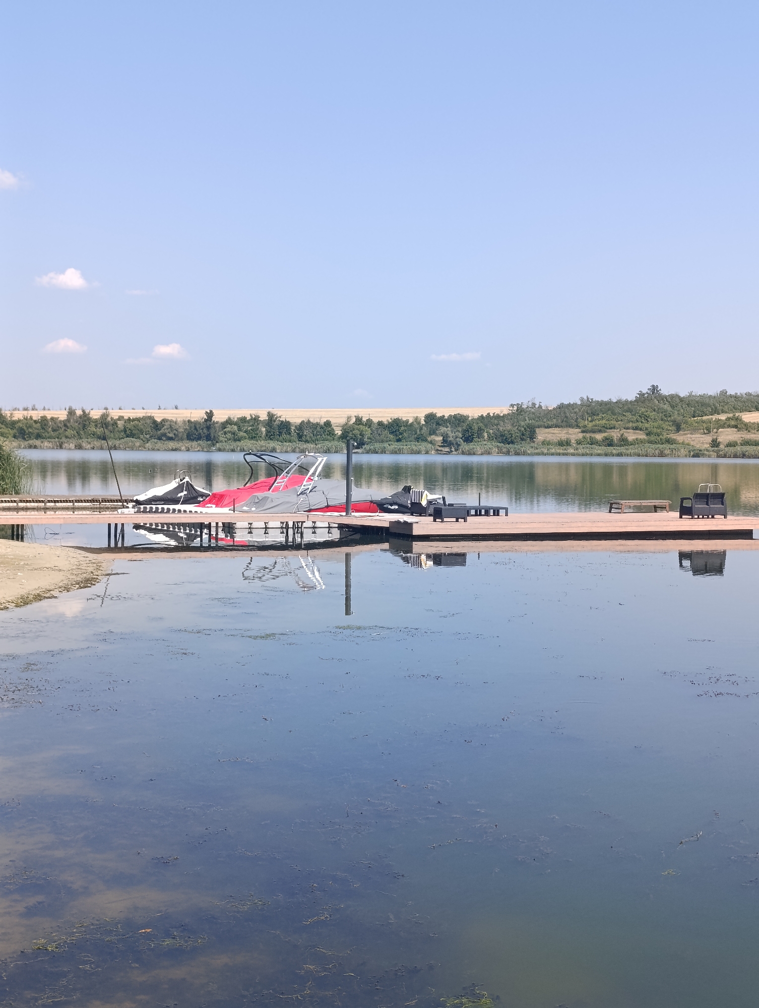 The image shows a wooden dock extending into a calm lake. On the dock, several covered boats, possibly jet skis or personal watercraft, are parked. The boats are covered with tarps, some red, some white, and some black. There are also black plastic chairs and a table on the dock. The lake is reflecting the sky and the objects on the dock. The background consists of a line of trees and a dry, grassy hill under a clear blue sky with a few wispy clouds. The location is the city of Bulatselivka, Ukraine.