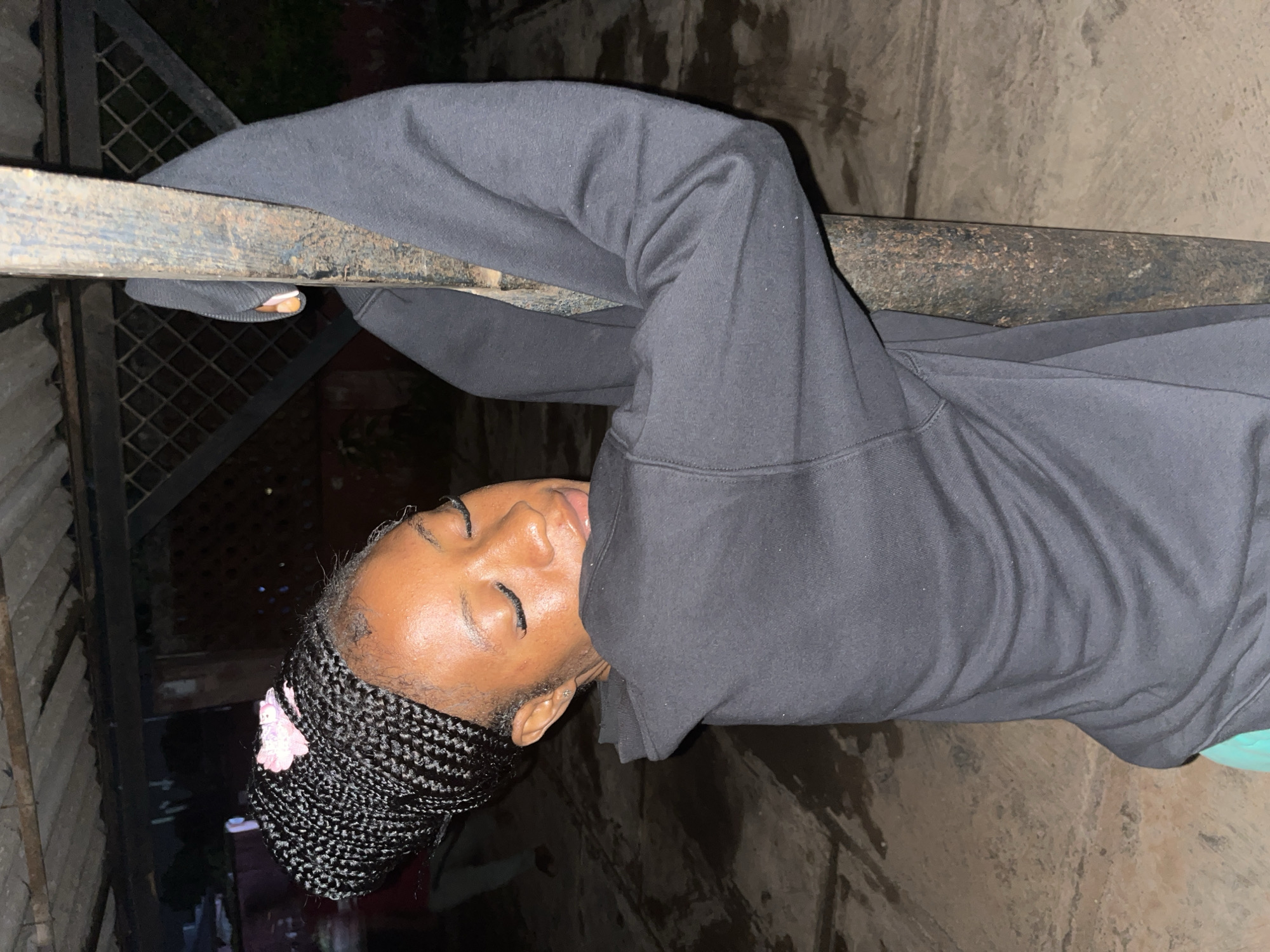 A young Black woman with braided hair and a pink accessory is leaning against a dark, rusty metal pole. She is wearing a black hoodie and green shorts. Her eyes are closed, and she appears to be resting her head on her arms which are wrapped around the pole. The background is dark and suggests an outdoor setting at night, possibly in a yard or open space with a concrete floor. The location is identified as Jos, Nigeria.