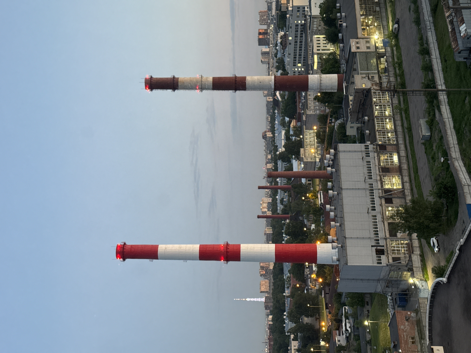 The image depicts an industrial and urban landscape in Saint Petersburg, Russia, observed during twilight conditions. Two prominent, vertically striped industrial chimneys, colored red and white, dominate the foreground. The left chimney features multiple red obstruction lights near its apex and midway point. The right chimney, slightly shorter and appearing older with more faded stripes, also displays multiple red obstruction lights.
At the base of the left chimney is a large, light-grey industrial building with numerous windows, suggesting a factory or power generation facility. Further to the right, another large, similar industrial building is visible, connected to a complex of smaller structures, fenced areas, and paved lots. Several vehicles, including a white SUV and a white bus, are parked within these industrial grounds. Scattered across the mid-ground are additional smaller, dark industrial smokestacks and various factory-like buildings.
Beyond the immediate industrial zone, a dense urban environment stretches into the distance, characterized by numerous multi-story buildings and significant tree cover. Streetlights and lights from within buildings are illuminated, indicating the low-light conditions. In the far distant left-center, the illuminated lattice structure of the Saint Petersburg TV Tower is clearly visible against the blue-grey sky. The scene is captured from an elevated vantage point, offering a panoramic view of the cityscape transitioning into night. No people are discernible in the image.