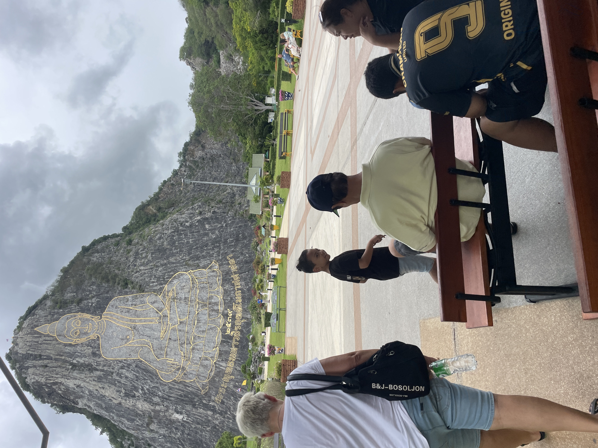 The image shows the Phra Tamnak Khao Chi Chan, a large Buddha image carved into a limestone cliff in Na Chom Thian, Thailand. The Buddha image is outlined in gold and is situated on a rocky mountainside. In the foreground, several people are visible. A woman in a white t-shirt and denim shorts walks away from the camera, carrying a black bag. A young boy in a black t-shirt looks back towards the camera. Further back, a group of people are seated on a wooden bench, facing away from the camera. The area appears to be a public space with paved pathways, benches, and landscaping. The sky is overcast with clouds.