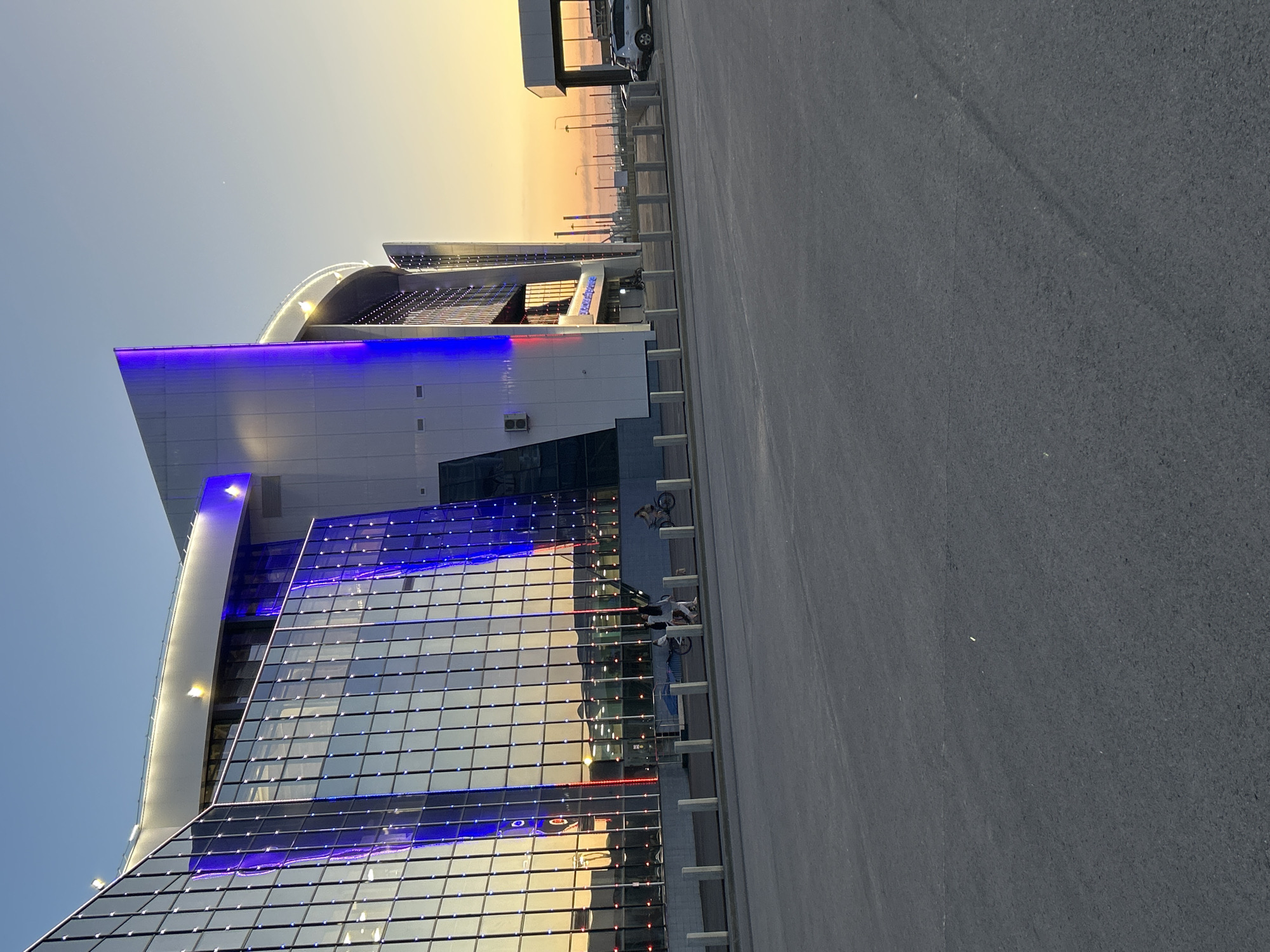 The image captures a modern building with glass facades and prominent blue lighting, possibly a sports arena or entertainment venue, in the city of Unknown, Unknown. The sky transitions from blue to orange, suggesting dusk or dawn. Two bicycles are parked near the building's entrance. A paved area occupies the foreground, indicating a parking lot or plaza. The building's design features curved elements and a combination of glass and solid panels. Street lights and parked vehicles are visible in the background, extending into the horizon.