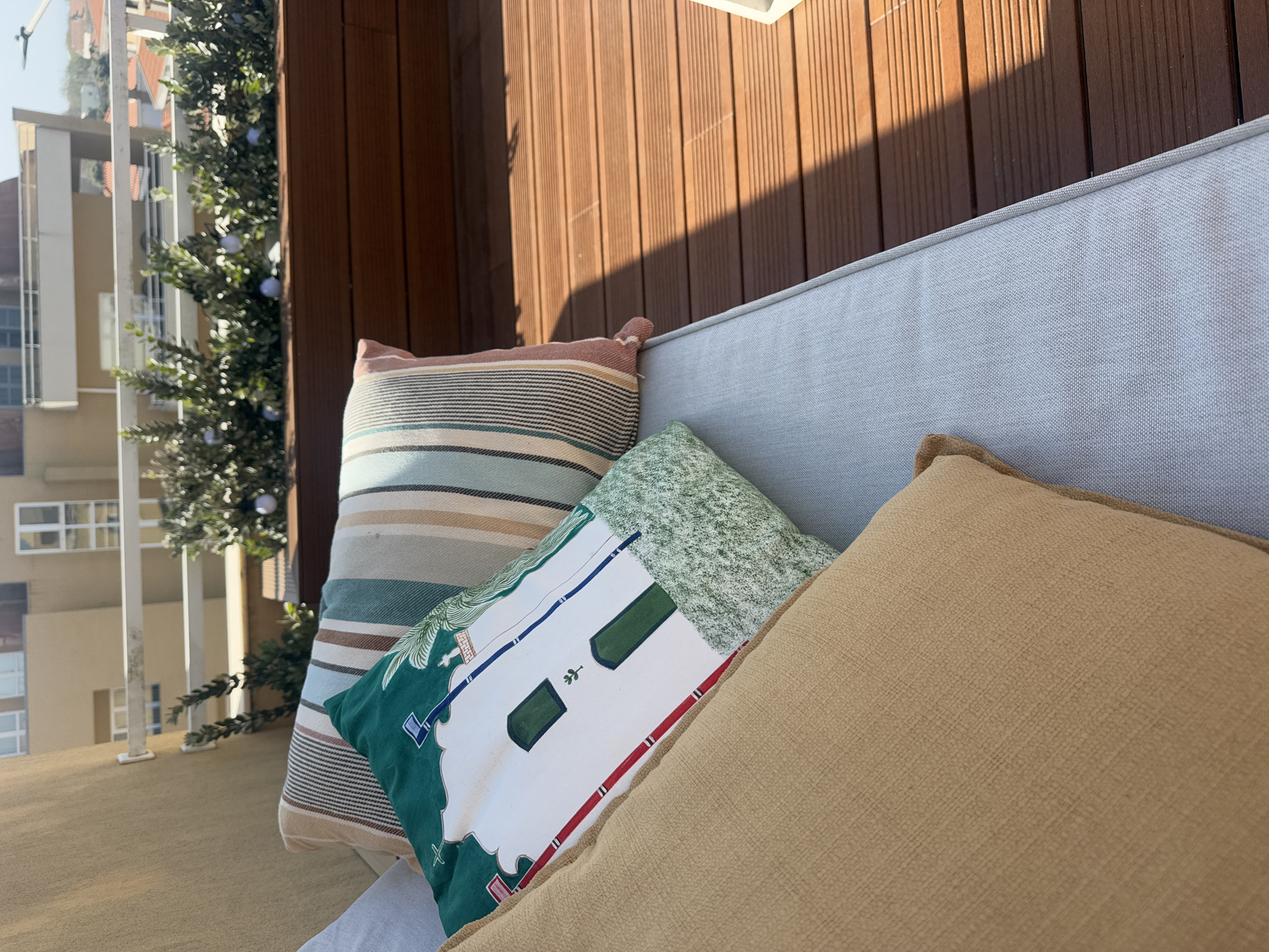 The image depicts a portion of a balcony, likely in Vila Nova de Gaia, Portugal, given the architectural style visible in the background. A wooden privacy screen forms the backdrop, partially illuminated by sunlight creating a sharp shadow. In the foreground, a light gray outdoor sofa is adorned with three cushions. The rearmost cushion features horizontal stripes in various earth tones and muted blues. A second cushion, with a primarily white base, has an illustration of a building with green accents. The foremost cushion is a solid tan color, its texture visibly coarse. The scene gives the impression of a comfortable and private outdoor space. Beyond the balcony's edge, buildings in a light neutral color can be seen, indicating an urban setting. A decorative green bush or small tree, ornamented with white spherical decorations, adds a touch of greenery.