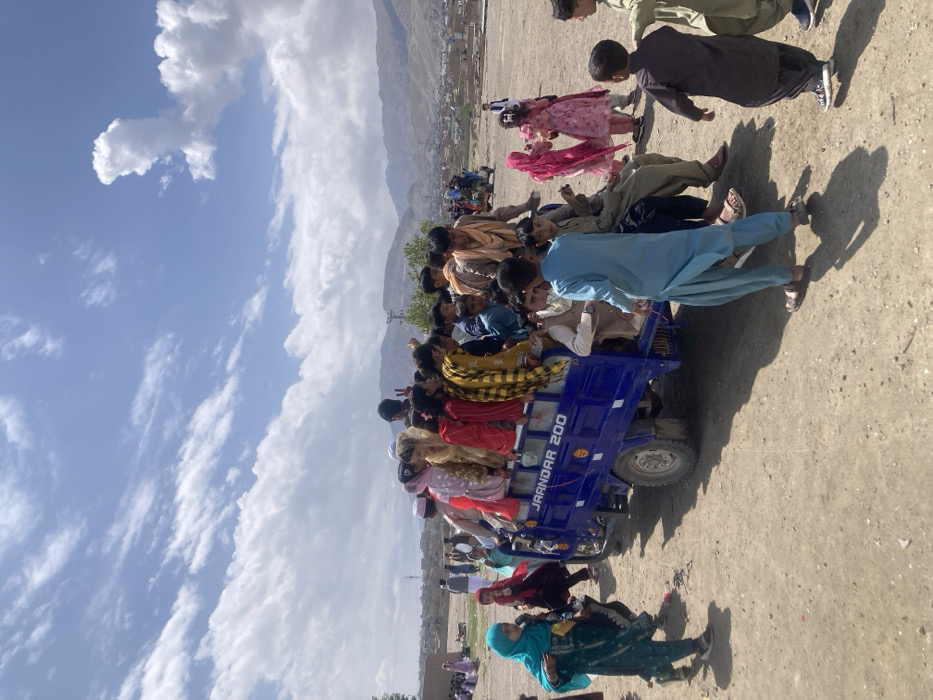 The image depicts an outdoor scene in Quetta City Tehsil, Pakistan, under a partly cloudy sky with prominent white cumulus clouds. A blue three-wheeled motorized vehicle, identifiable by "JAANDAR 200" lettering on its cargo bed, is centrally positioned on a barren, dirt ground surface. The vehicle's open cargo bed is densely populated with approximately 15 to 20 individuals, predominantly children and young adults, who are either seated or standing. They wear various traditional garments, including shalwar kameez, in diverse colors; one individual in the rear of the vehicle displays a "peace" hand gesture. Numerous other children are present on the ground adjacent to the vehicle, some appearing to be in motion. In the left foreground, a young girl in a teal shalwar kameez and matching head covering walks towards the left. A boy in a light blue shalwar kameez stands near the vehicle's right side, facing away. Additional figures and scattered residential buildings are visible in the distant background, with a range of mountains beyond.