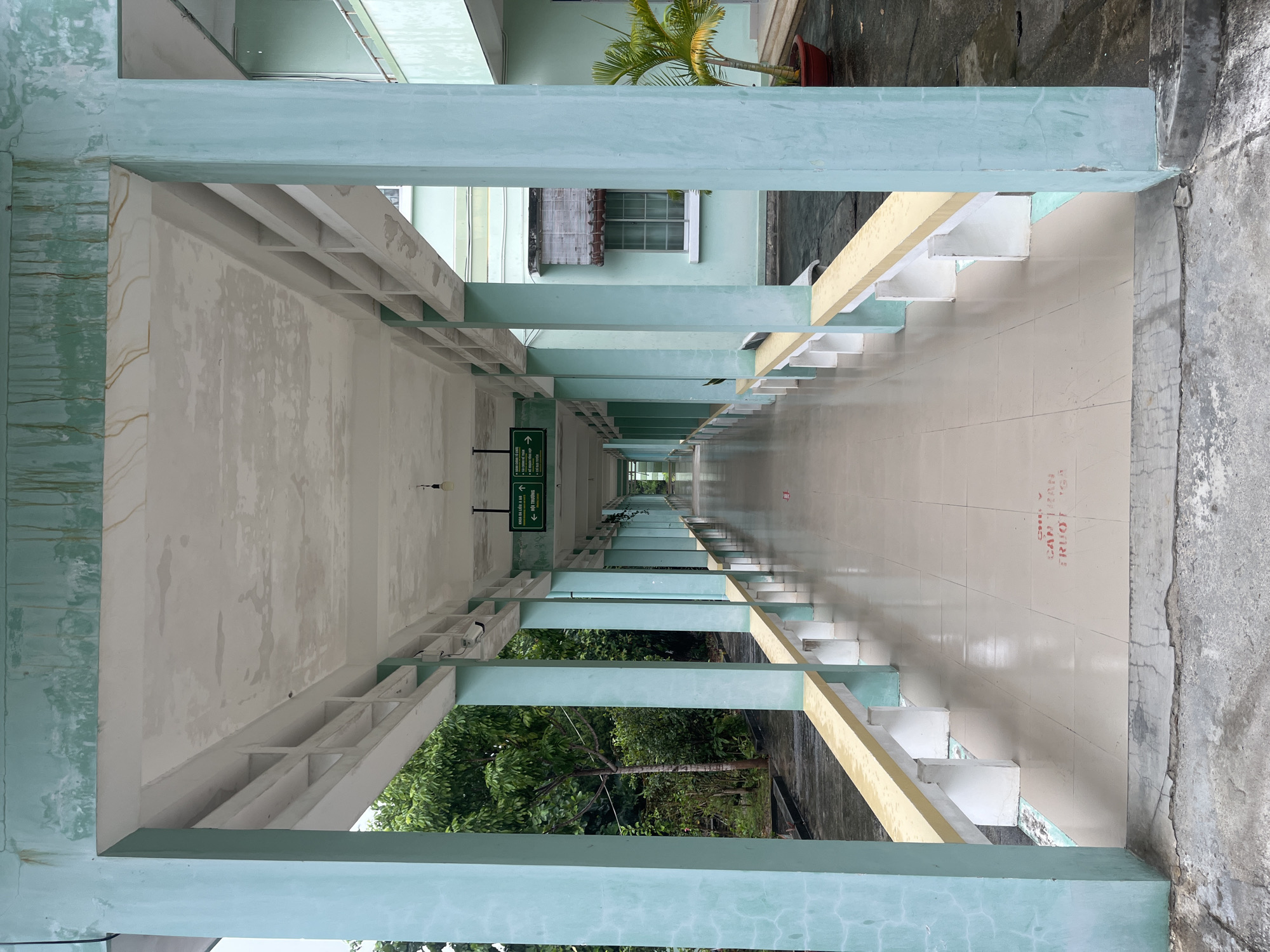 The image depicts a long, open-air corridor in what appears to be a building located in Bac Nha Trang Ward, Vietnam. The corridor is defined by a series of light blue concrete pillars and beams supporting a white ceiling. The floor is tiled in light beige. On one side of the corridor is a series of stepped platforms painted with a yellow stripe, leading down to an outdoor area. Green foliage is visible on both sides of the corridor, suggesting the presence of trees and vegetation. Signage is visible hanging from the ceiling, and the overall impression is of a weathered, utilitarian structure. The perspective is from one end of the corridor, looking lengthwise.