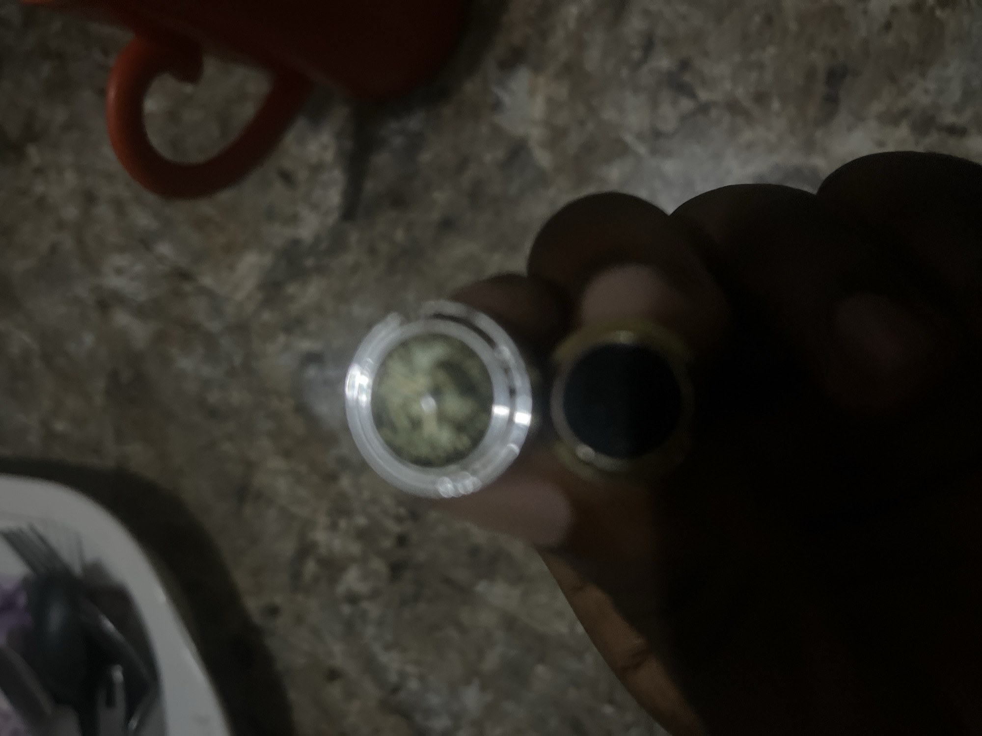The image shows a person's hand holding two rings, one on top of the other. The top ring is silver and contains a substance that appears to be green plant material. The bottom ring is black with a gold band. The background is a mottled brown surface. There is an out-of-focus red mug and a plate with utensils in the upper left corner. The scene is dimly lit, suggesting it is indoors. The image was taken in Jalingo, Nigeria.