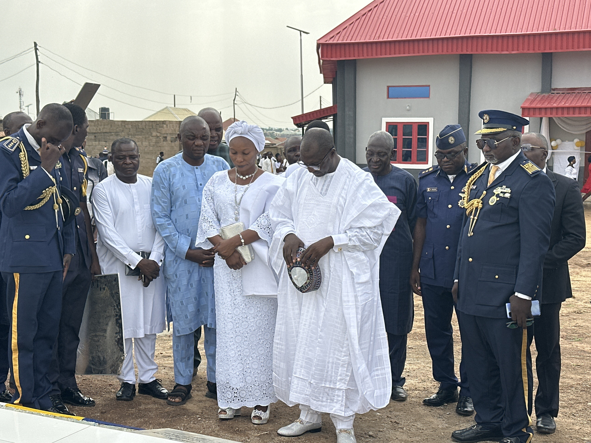 A group of people are gathered outdoors in what appears to be a solemn event in Goni Gara, Nigeria.
The group consists of men and one woman, of African descent, dressed in a mix of traditional and formal attire. Several men are wearing Nigerian Air Force uniforms. One man is dabbing his eye, suggesting he is experiencing grief. A woman in white lace stands next to a man in a white robe. He is holding a patterned bag. The other men are dressed in traditional Nigerian attire or formal suits. A picture frame leans against a man's leg.
In the background is a building with a red roof. The sky is overcast.