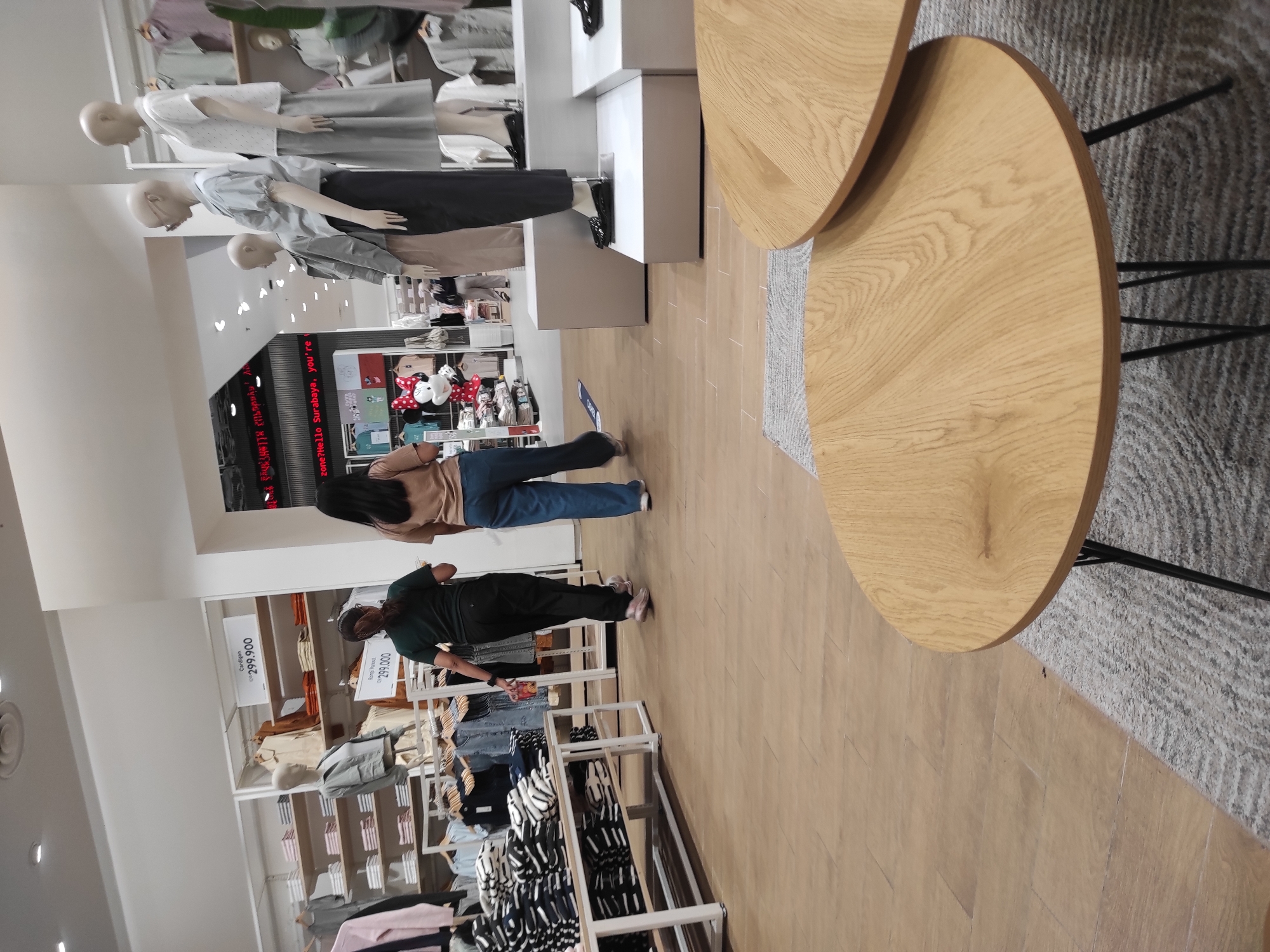The image shows an interior view of a clothing store, likely in Surabaya, Indonesia, based on the signage visible in the background which reads "zone? Hello Surabaya, you're". In the foreground, two wooden coffee tables with black metal legs are positioned on a grey rug. In the middle ground, two women are walking through the store. The woman on the left is wearing a dark green t-shirt and black pants, while the woman on the right is wearing a light brown t-shirt and blue jeans. The store features several clothing racks filled with garments, shelves stocked with folded clothing, and mannequins displaying outfits. A large poster of Minnie Mouse in a red polka dot dress is also visible on a wall in the background. The store appears well-lit and organized, with a focus on casual wear.