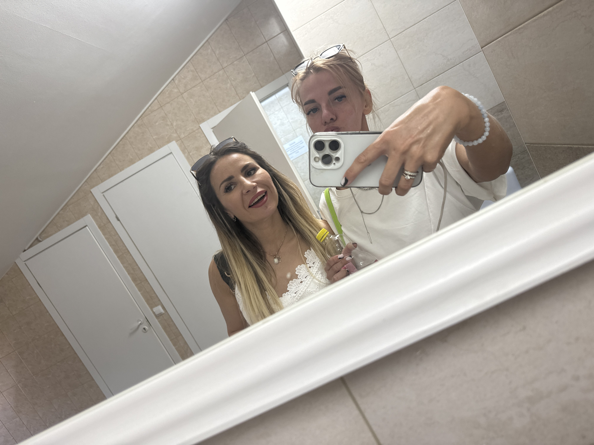 In Saint Petersburg, Russia, a selfie is taken in a brightly lit, tiled restroom. Two women are reflected in a large mirror that spans across the frame. The woman in the foreground, on the left, has long blonde hair with streaks of brown and is wearing a white lace top and sunglasses perched on her head. She holds a small yellow bottle in her hand. The woman in the background, on the right, has blonde hair pulled back and is wearing a white top. She is holding a smartphone in her right hand, capturing the reflection. The restroom features beige tiled walls and two white doors are visible in the background. A white framed mirror dominates the foreground, with its reflection stretching across the scene.