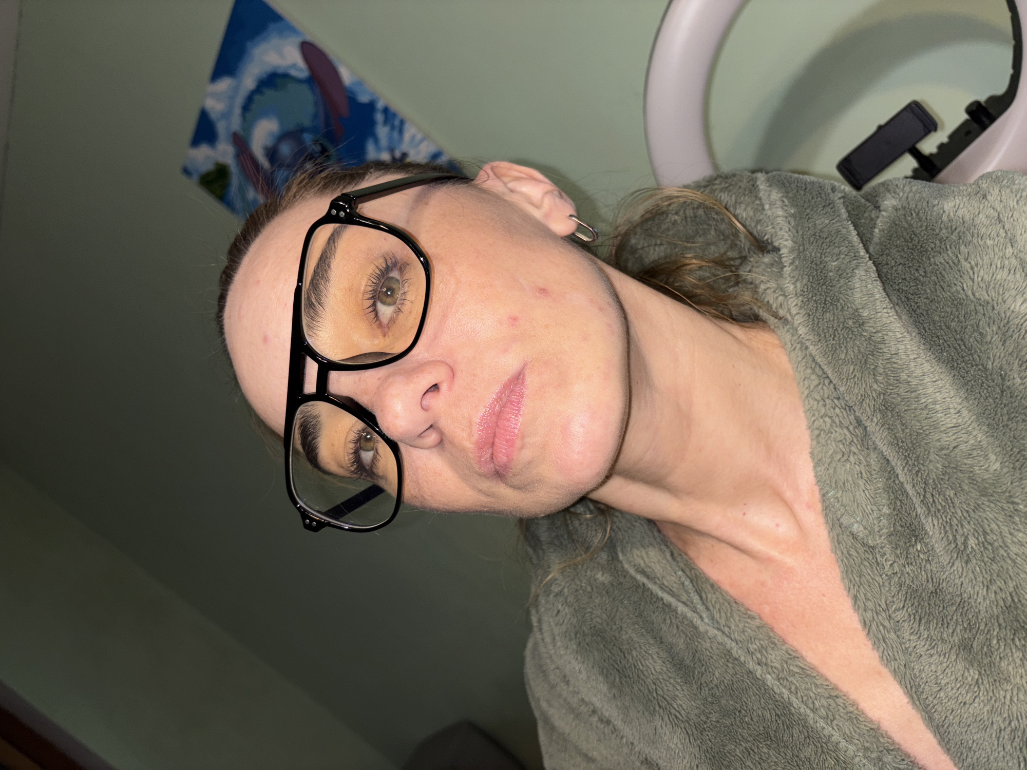A medium close-up, eye-level, selfie-style shot shows a woman with light skin and auburn hair, wearing large black-rimmed glasses and a fuzzy olive-green robe. She is looking directly at the camera with her head tilted slightly. She has light makeup on, including eyeshadow and lip gloss. Her skin shows some redness and blemishes on her cheek. A silver hoop earring is visible in her right ear. In the background, the wall is a pale green, with a painting hanging on it that appears to depict a blue wave with red abstract shapes. A portion of a light-colored, possibly medical, device with a black handle is visible on the right side of the frame. The lighting is dim, casting a slight shadow on her face. The image appears to have been taken indoors.
