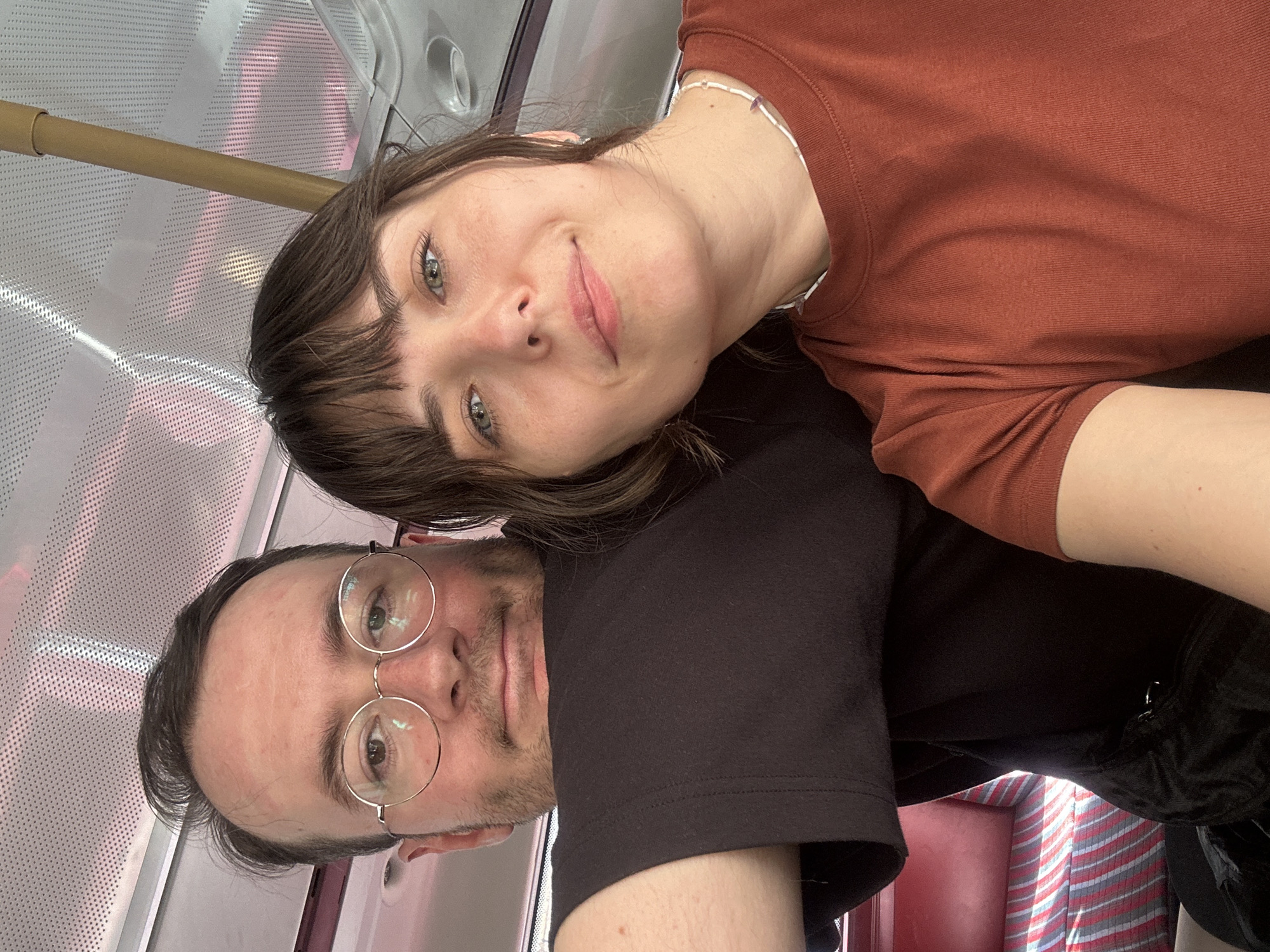 A man and a woman are taking a selfie on what appears to be a bus. The man is wearing a black t-shirt and round glasses, and the woman is wearing an orange t-shirt and a beaded necklace. They are seated next to each other, with the man's head resting on the woman's shoulder. The background shows the interior of a bus, with red and white striped seats visible. The ceiling of the bus is white and perforated. There is a yellow pole extending from the ceiling down towards the right side of the frame. The lighting suggests it is daytime.