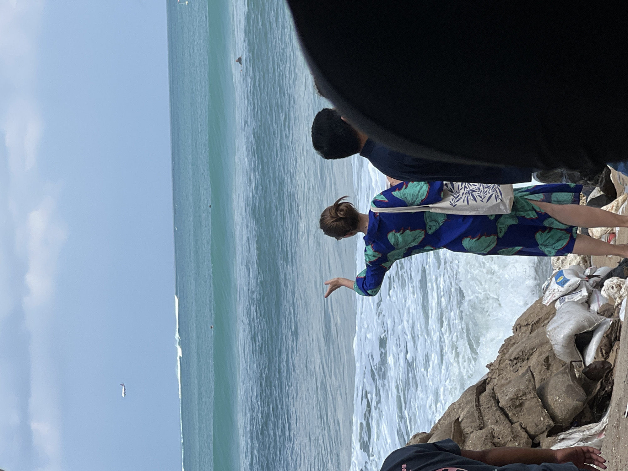 An outdoor, eye-level shot captures a wide view of the ocean and beach in Kuta, Indonesia. In the distance, an airplane is visible against a cloudy sky. The ocean is a vibrant turquoise, with visible waves and a few surfers in the water. In the foreground, three people stand near a rocky embankment and some piled-up debris. A woman in a blue dress with a green leaf print faces away from the camera, gesturing towards the ocean with her right hand. She carries a white tote bag with a blue pattern. Beside her, a man in a dark shirt also faces the ocean. The edge of a fourth person, wearing black clothing, is visible in the far right of the frame. The beach appears to be in a state of disarray, with rocks, sand, and various types of trash scattered along the shoreline.