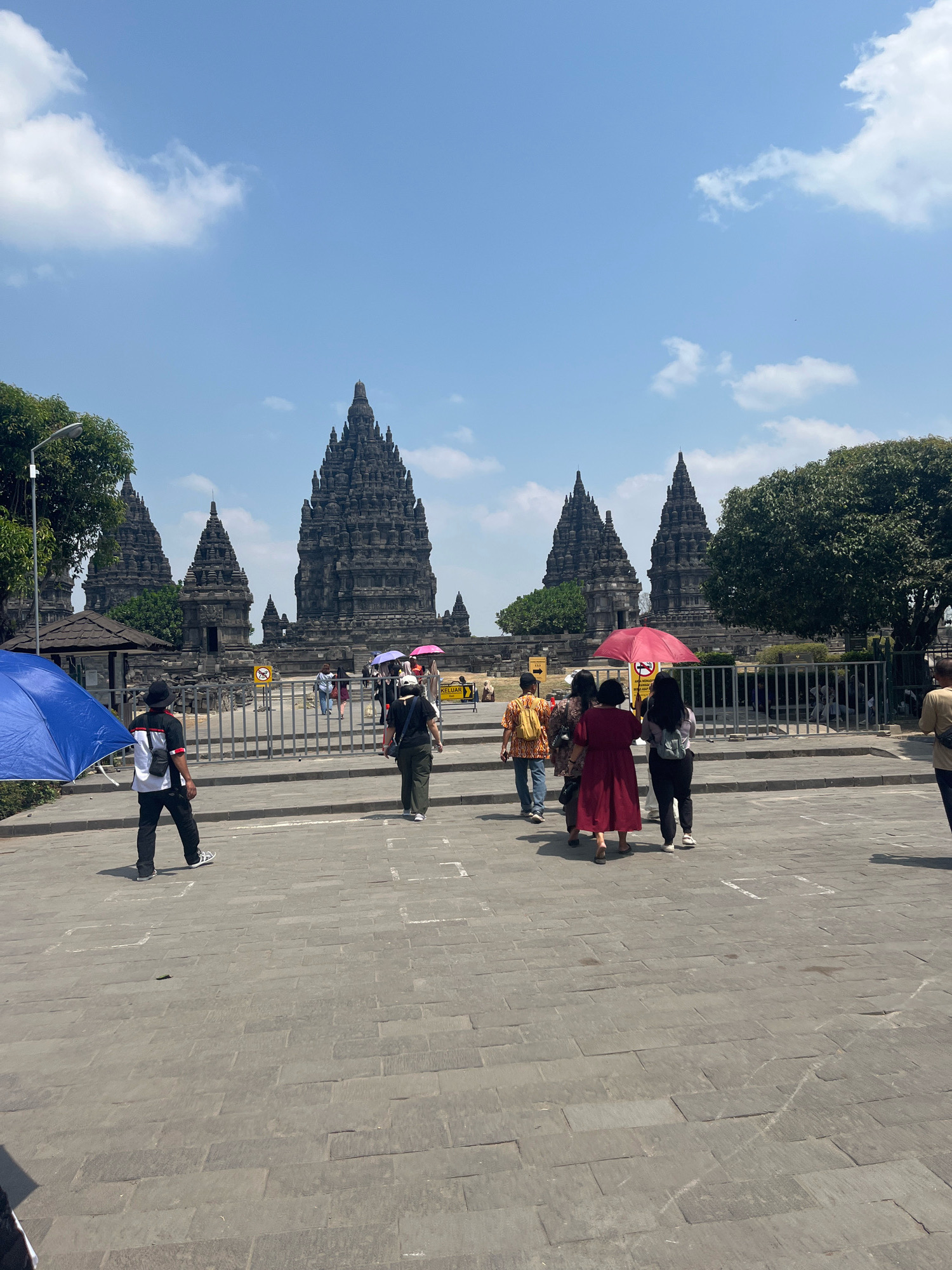 The image depicts an outdoor scene at Prambanan Temple, a 9th-century Hindu temple complex located in Bokoharjo, Sleman, Special Region of Yogyakarta, Indonesia. Several people are visible walking on a paved area in front of the temple complex, which features a series of tall, intricately carved stone towers under a bright blue sky with scattered clouds.
In the foreground, on the left, a person in a black hat, black and white shirt, and black pants walks away from the camera. A large, bright blue umbrella is open to the left of this individual, casting a shadow. Further back, several other individuals are walking towards the temple. One person is wearing a red dress and carrying a red umbrella. Another person is wearing a dark top and olive green pants. A group of people are walking in a cluster.
The temple structures themselves are large and made of dark stone, with multiple tiered roofs and pointed spires. They are set behind a metal fence. Trees with lush green foliage are visible on the sides and behind the temple structures. The ground is paved with large, rectangular stone slabs. Signs with yellow and black markings are visible near the entrance to the temple grounds. The overall impression is of a sunny day with tourists visiting a significant historical and religious site.