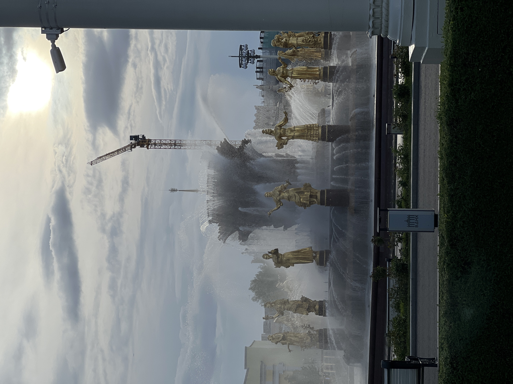 The image shows the Friendship of Nations Fountain at the Exhibition of Achievements of National Economy (VDNKh) in Moscow, Russia. Golden statues representing different Soviet republics stand amidst jets of water. In the background, a construction crane is visible against a cloudy sky. A surveillance camera is seen in the top left corner. A sign with the VDNKh logo is present on the right side of the frame, next to a hedge.