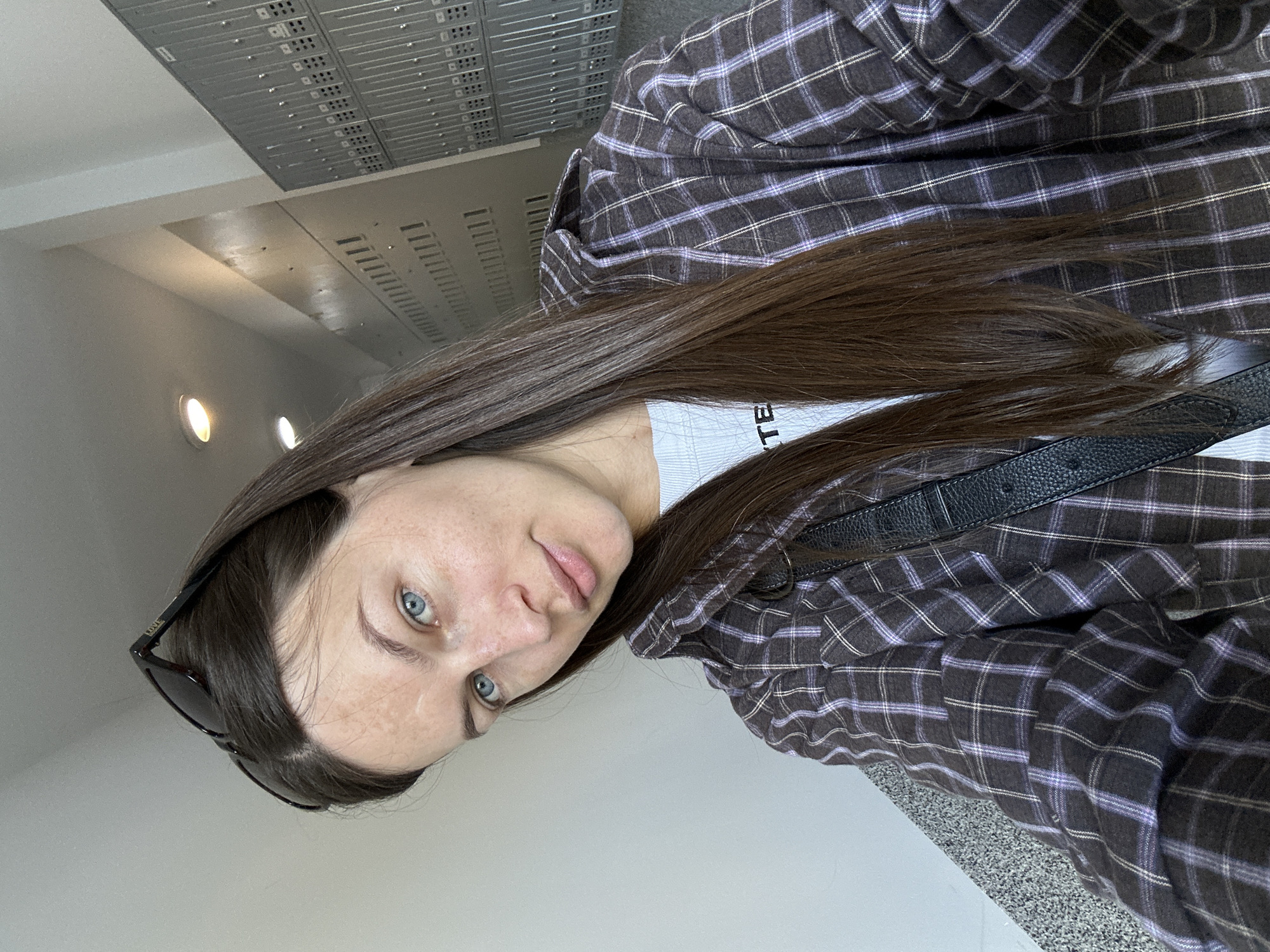 A woman is looking at the camera. She has long brown hair, fair skin, and light blue eyes. She is wearing a plaid shirt and a black bag strap is visible across her body. Sunglasses are perched on her hair. The background is a nondescript interior space, possibly a hallway, with light fixtures and metal panels on the ceiling. The location and city of Budva, Montenegro, cannot be determined from the image.