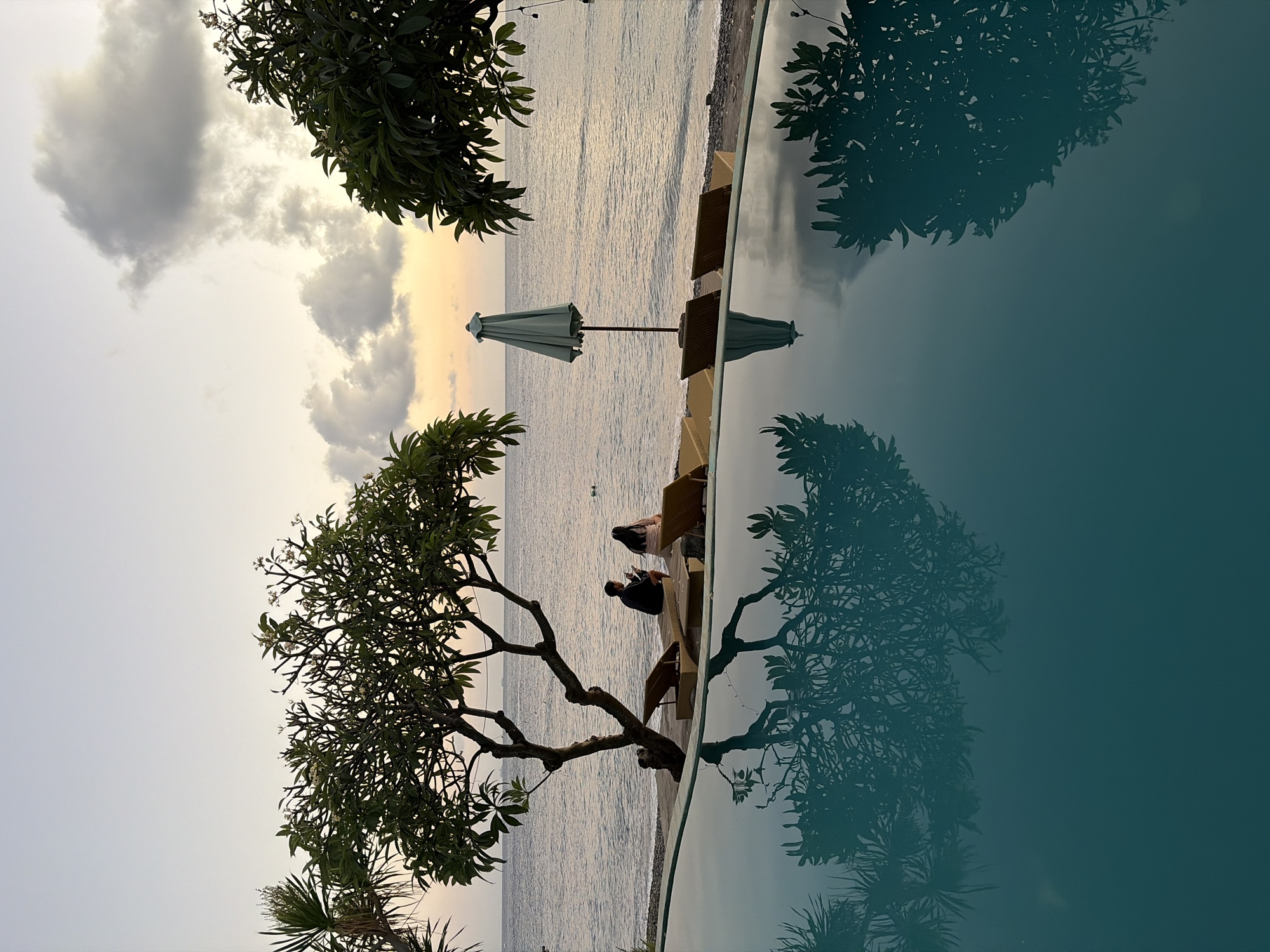 The image depicts a serene scene by the sea in Abang, Indonesia, likely at dusk. In the foreground, a tranquil swimming pool reflects the surrounding environment. Beyond the pool, a beach is lined with lounge chairs and trees. Two individuals are seated on lounge chairs near the water's edge, facing the sea. One appears to be looking at a mobile device. An umbrella is open between two lounge chairs. The sea stretches to the horizon, where the sky transitions through soft hues of orange and gray, indicating the setting sun. Clouds are present in the sky. The overall atmosphere is peaceful and relaxing, characteristic of a tropical resort setting.