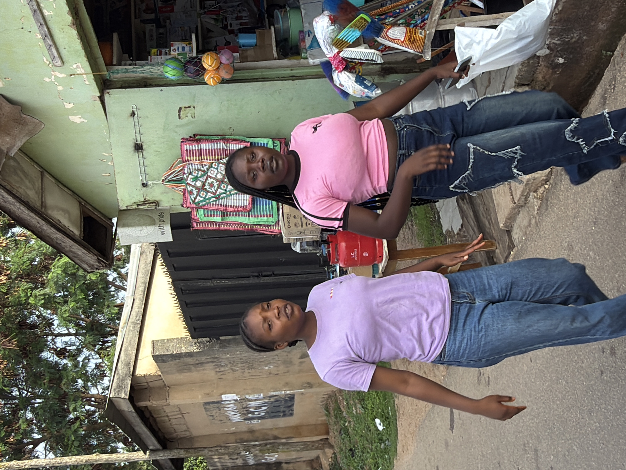 The image depicts a daytime street scene in Jos, Nigeria, featuring two young adult women walking on a paved pathway alongside a commercial building.
On the left, a light brown, weathered wall constructed from concrete blocks displays multiple faded black posters or graffiti, including visible letters "GCM." Above this wall, a dark metal gate with sharp, pointed anti-climb elements is partially visible, topped by a corrugated metal roof. In the upper left and central background, dense green foliage of large trees is visible against a light blue sky.
The woman on the left wears a lavender short-sleeved t-shirt with a small red and white striped emblem on the chest and faded blue straight-leg jeans. Her dark hair is styled in tight braids pulled back from her face. Her mouth is slightly open as if speaking, and she looks forward.
The woman on the right wears a pink short-sleeved crop top with black stripes on the sleeves and a black silhouette logo on the chest. Her dark hair is styled in long, thin braids. She wears dark blue jeans featuring distinctive star-shaped cutouts with frayed edges along the legs. She carries a white plastic shopping bag and a dark-colored smartphone in her right hand. Her left arm is slightly extended. Both women are walking from right to left across the frame on a worn asphalt or concrete pathway, bordered by patches of green vegetation.
On the right side of the image, a single-story commercial building, likely a shop or stall, is visible. Its exterior is painted a peeling light green, revealing lighter paint layers underneath. It has a weathered corrugated metal roof with visible wooden support beams. Near the open entrance, two red propane gas cylinders, one marked "LC," are placed on a small wooden table. Several brightly colored woven bags or mats with geometric patterns are hanging on a metal rack near the entrance. A string of spherical objects (green, purple, orange) is also suspended nearby. Inside the shop, various household goods, including plastic containers and packaged items, are stacked on shelves. On the far right, cleaning brushes and other plastic items are displayed vertically.