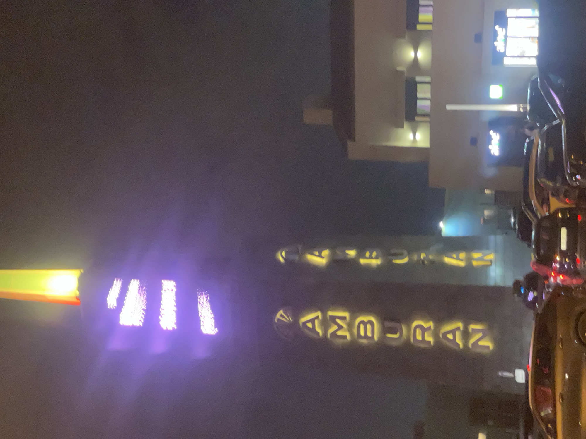 The image depicts an outdoor scene at night, focusing on two tall, illuminated structures. The left structure has vertical text in glowing yellow letters spelling "TAMBURAN". Above the yellow text, there is a section illuminated with purple light, and the very top of this structure is lit with bright yellow and red light, resembling a flame.
To the right of the "TAMBURAN" structure is another similar, taller structure, also illuminated vertically with yellow letters. The visible letters appear to spell "FUAN".
In the background, a modern building with multiple windows and external lights is visible. Some of the windows have colored lights. Two signs reading "aleef" are visible on the ground floor of this building.
The foreground is occupied by several cars, suggesting a street or parking area. The lighting indicates it is nighttime, and there is some atmospheric haze or light diffusion, particularly around the illuminated elements. The overall scene appears to be in an urban environment, possibly a public space or entertainment area. The presence of the Sabunchu Raion, Azerbaijan is indicated in the prompt, placing the scene in that specific location.