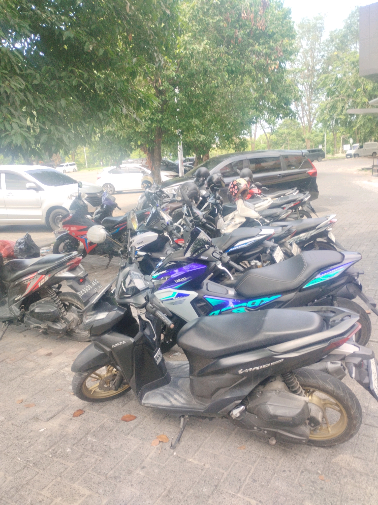 The image depicts a parking lot in Percut Sei Tuan, Indonesia, filled with numerous parked motorcycles. The scene is dominated by a variety of scooter-style motorcycles, with a black Honda scooter prominently featured in the foreground. Other motorcycles, including a red and black Yamaha, a blue and black Yamaha, and a white and blue Yamaha, are visible behind it. Several helmets are placed on the handlebars or seats of the motorcycles. In the background, a black SUV and white cars are parked, suggesting a public area or commercial establishment. The ground is paved with interlocking concrete blocks, and several trees with dense green foliage provide shade over the parking area. The lighting suggests it is daytime, possibly overcast or late afternoon, with soft shadows.
