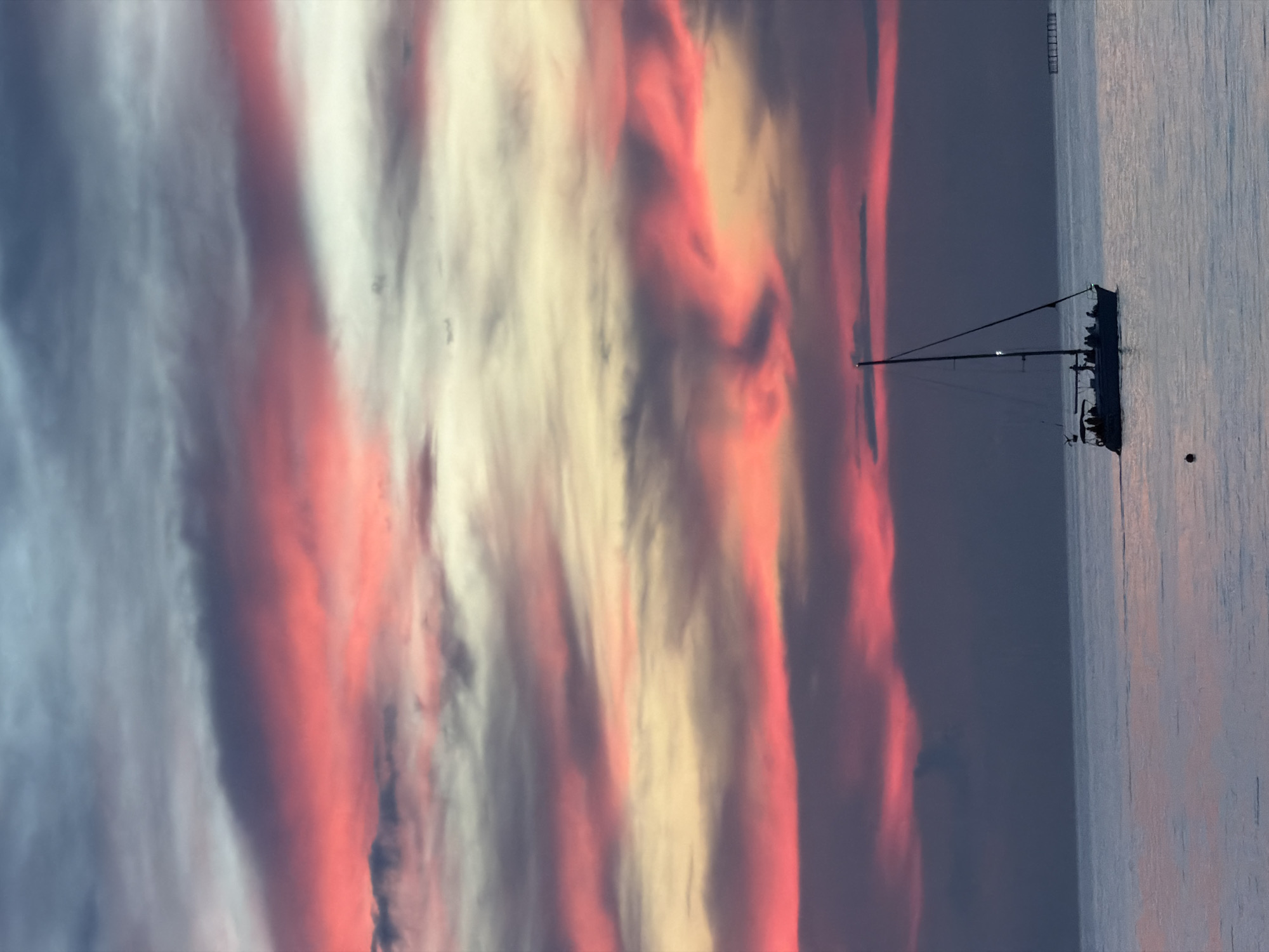 A sailboat with people onboard is sailing on calm water during sunset. The sky is filled with colorful clouds in shades of pink, red, yellow, and blue. In the distance, there appears to be a structure on the water, possibly a pier or a fishing platform, on the right side of the image. The scene suggests a tranquil evening on the coast. The location is the coast of Sochi, Russia.