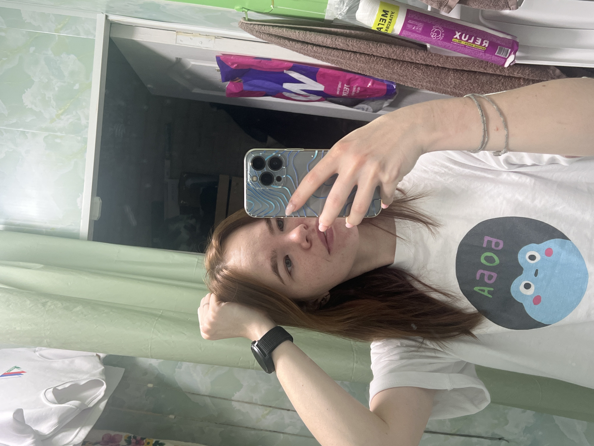 The image shows a young woman taking a selfie in a bathroom. She is holding a smartphone with a patterned case, partially obscuring her face. She wears a white t-shirt with a frog design and a black smartwatch on her left wrist. Her hair is brown. The background includes a bathroom mirror, green and white tiled walls, towels, and toiletries. Based on the "RELAX" text on the packaging, the image is likely from the city of Chaikovsky, Russia.
