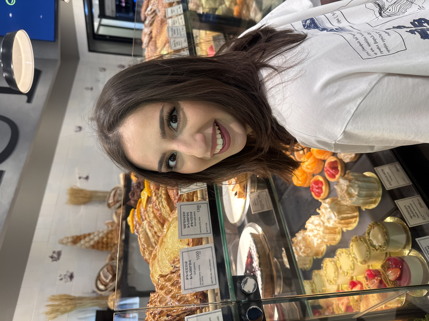 A young woman with long, dark hair and a bright smile is pictured inside a well-lit bakery or pastry shop in Yerevan, Armenia. She is wearing a white t-shirt with a blue and white graphic design that includes some indistinct text. Her head is tilted, and she is looking directly at the camera.
She stands in front of a glass display case overflowing with a diverse array of baked goods and desserts. Among the visible items are large golden-brown pastries, resembling apple tarts, with price tags in both Armenian and English. Specific visible text on the tags includes "ԹԽՎԱԾՔ ԽՆՁՈՐՈՎ Grill pomme 1000" and "ԿՏՐԱՏՎԱԾ ԽՆՁՈՐՈՎ ԹԽՎԱԾՔ Sliced apple pastry 1300", indicating prices in Armenian drams. Other desserts include small tarts with red fruit toppings, orange-colored pastries, white-frosted confections, and possibly meringues or miniature cakes. Decorative elements resembling sheaves of wheat are also seen in the background of the display. The woman's joyful expression suggests she is enjoying her visit to the bakery.