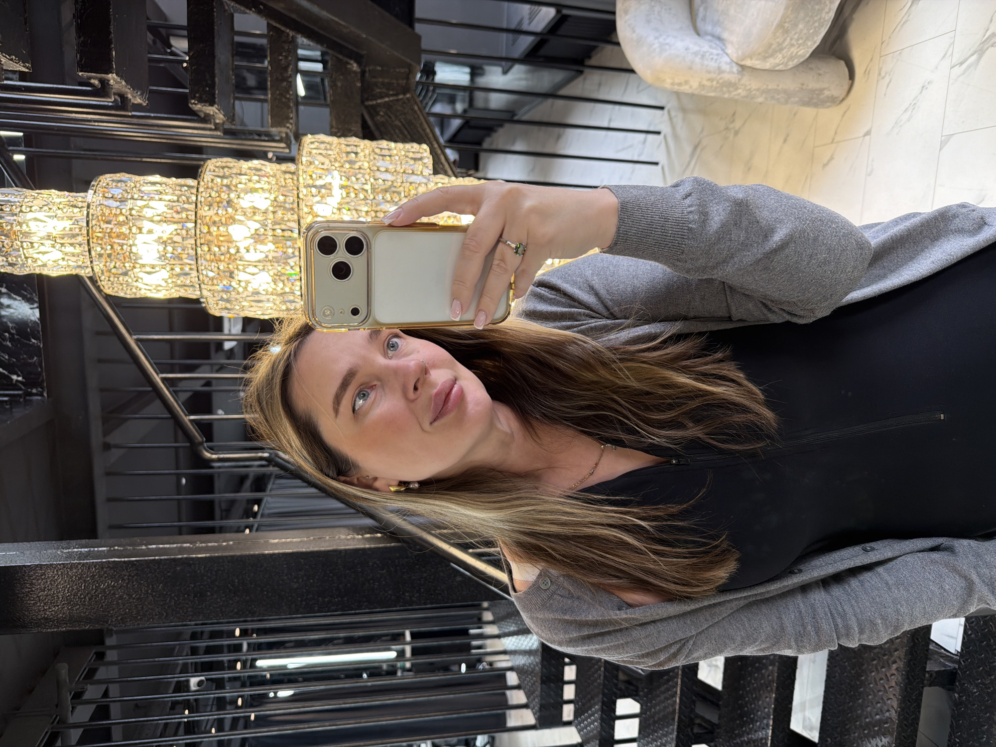 The image is a rotated selfie taken indoors. A Caucasian woman with long brown hair highlighted blonde stands facing the camera, holding up a cell phone with a gold-colored case in her right hand. She wears a black garment under a gray cardigan, with a gold chain around her neck and small gold earrings. Her ring finger sports a ring with what appears to be a green gemstone.
Behind the woman is a modern chandelier consisting of several tiers of clear, glittering cylindrical shapes. The backdrop features elements of modern architecture, including a black staircase with metal bars and metal grating steps. A white decorative sculpture is visible in the background. The image does not provide enough detail to definitively pinpoint the exact location in Moscow, Russia.