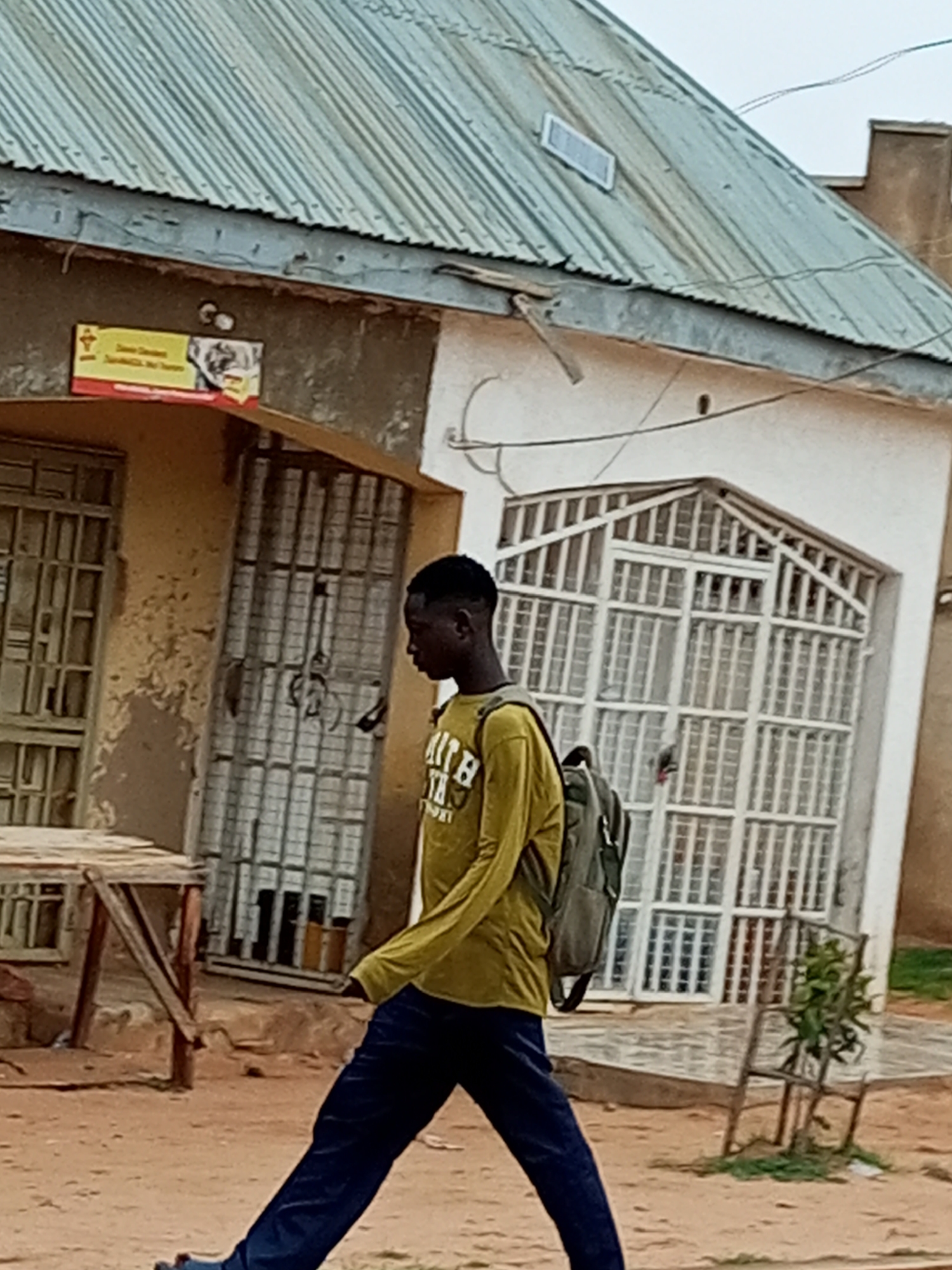 A young Black male with short hair is walking from right to left in front of a building in Gombe, Nigeria. He is wearing a long-sleeved yellow shirt with writing on it and dark blue pants, and he has a gray backpack slung over his right shoulder. The building appears to be made of concrete with a corrugated metal roof. There are barred windows and doors on the building, and a sign with yellow and red text is affixed to the wall to the left. The ground is dirt and sand. The weather appears overcast.