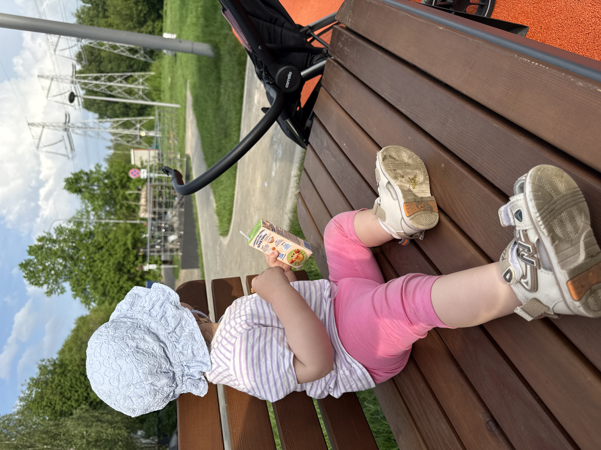A child wearing a blue hat, a striped shirt, and pink leggings is lying on a wooden bench while drinking from a juice box. The bench is next to a black baby stroller. In the background, there are green trees and electrical power lines. The location is in Moscow, Russia.