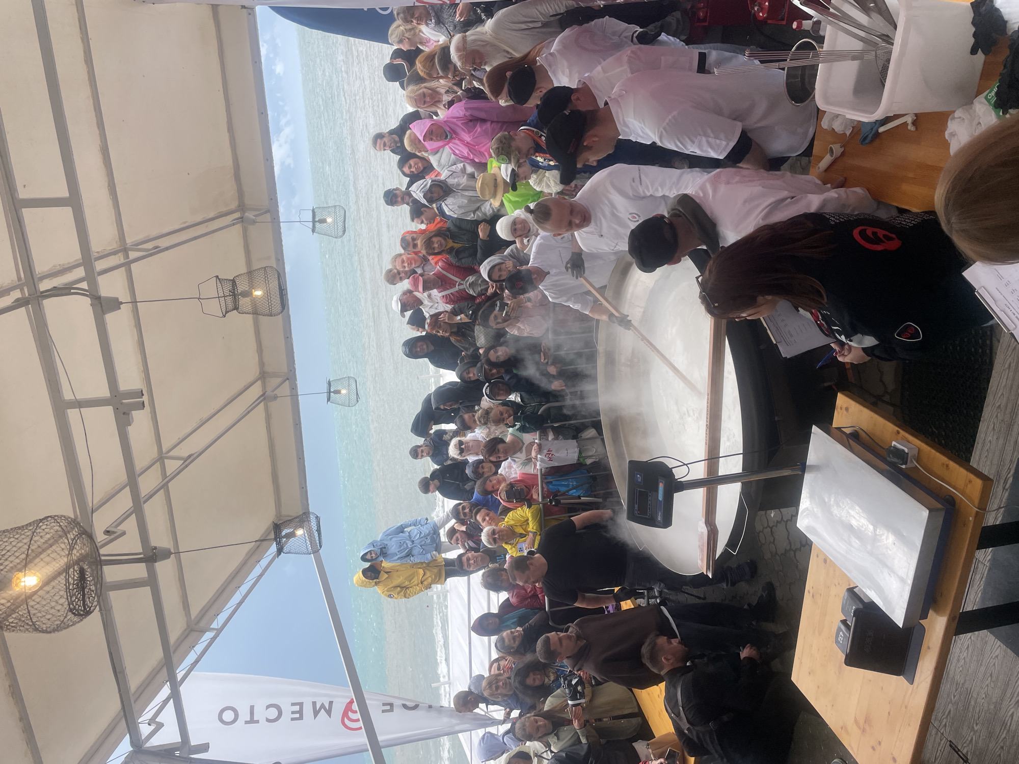 The image captures a large crowd gathered under a tent near the sea, possibly in Sochi, Russia, based on the presence of Russian script in the background. The event appears to center around a massive pot, filled with a steaming substance, likely food, with chefs in white uniforms stirring it. The crowd is diverse, with people of varying ages and attires. Some individuals are taking notes, possibly judging or documenting the cooking process. Tables with equipment are present, indicating a cooking demonstration or competition. The backdrop features the sea and sky, partially obscured by the tent. Overhead lighting fixtures are also visible. The overall scene suggests a public cooking event, possibly aimed at setting a record or promoting local cuisine.