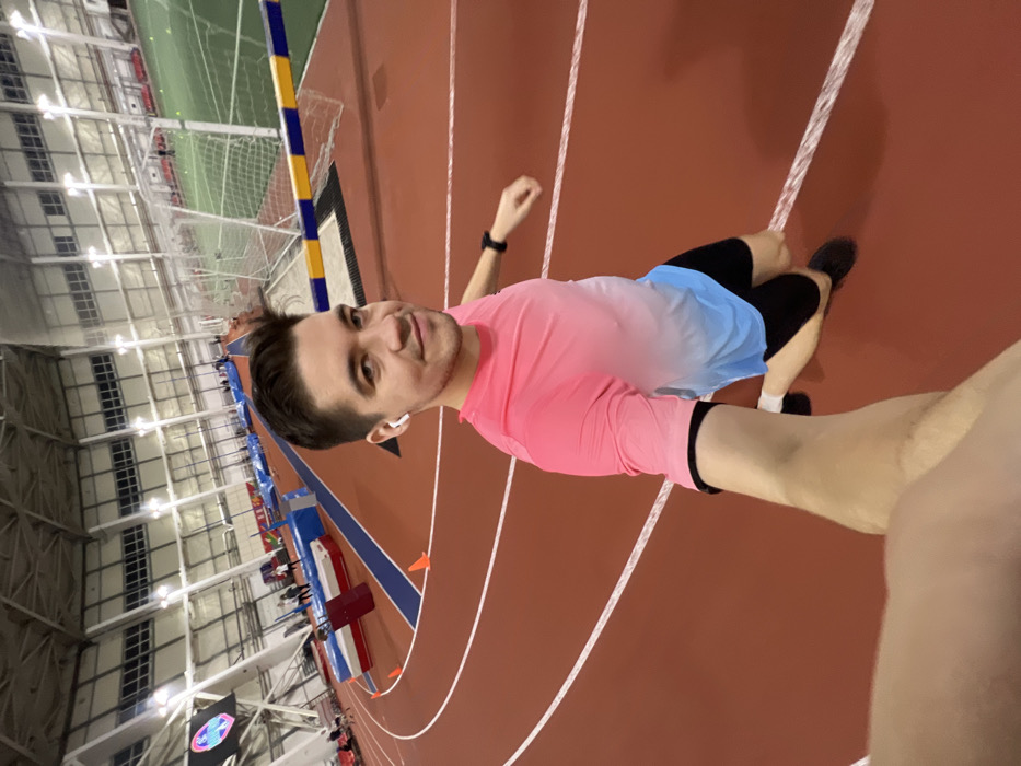 A man takes a selfie indoors at a track and field facility. He wears a pink and blue ombre shirt, black shorts, and black sneakers. An athletic track in shades of brown is visible beneath him. The track has white lines painted on it. Behind the man are rows of seating, hurdles, and a net. The ceiling is high and industrial with numerous lights. He wears a smartwatch and an earbud.