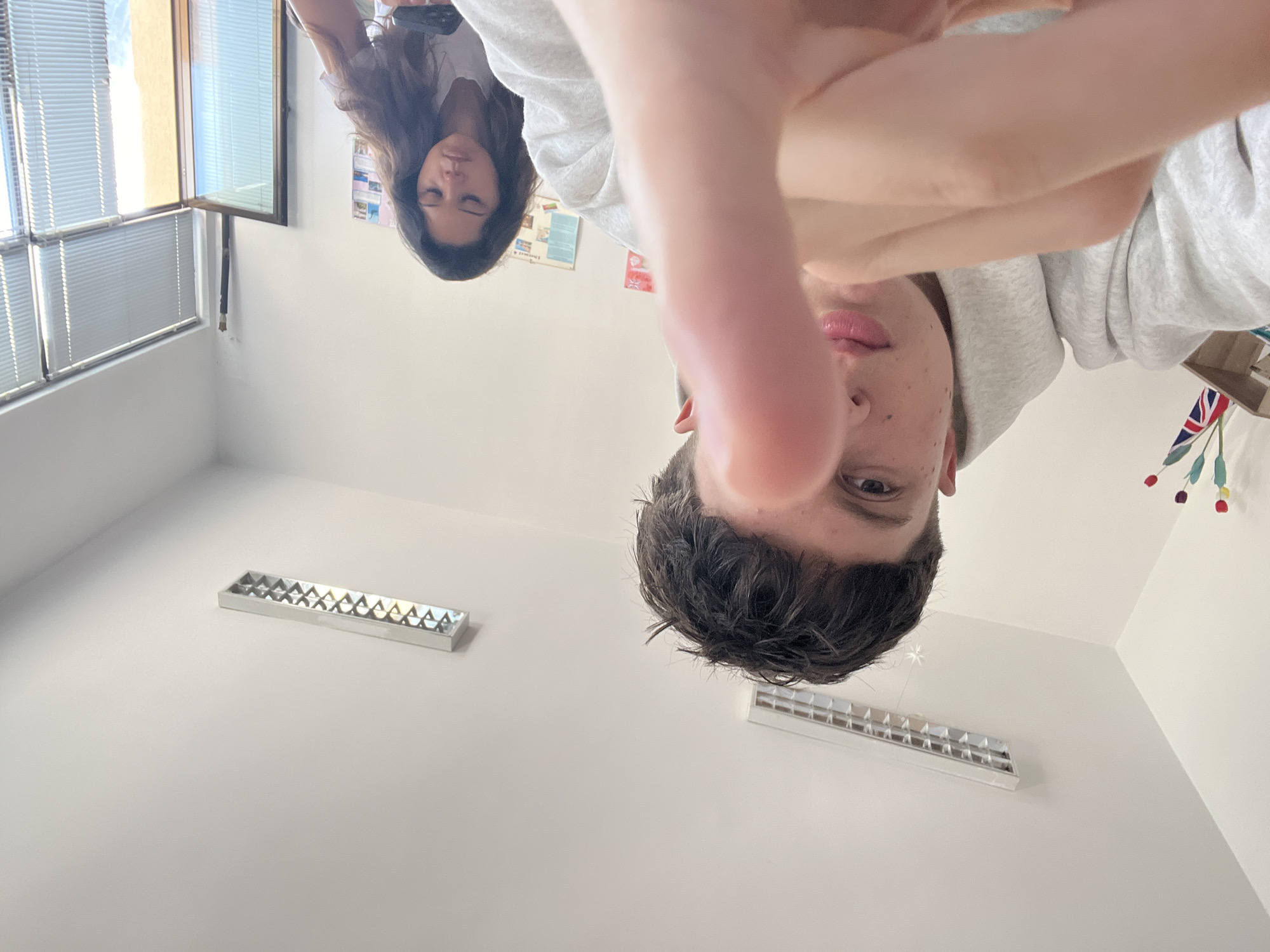This interior photograph, taken in Zlatograd, Bulgaria, presents an unusual perspective, as if captured from a low angle looking upwards, and then rotated 90 degrees clockwise. This rotation makes the subjects and the room appear to defy gravity.
In the immediate foreground, a young male's face, sporting dark, messy hair and visible freckles, is partially obscured by his extended hand. He wears a grey hooded sweatshirt, and his right eye is open, looking directly at the camera. Behind him and slightly to his left, a female with long, dark hair rests her head, her eyes closed, suggesting a moment of calm or introspection. She is wearing a light-colored top.
The room features light-colored walls and a ceiling with two rectangular fluorescent light fixtures. To the left, a window fitted with horizontal venetian blinds allows bright daylight to illuminate the space. A black pipe or conduit descends from the window frame. On the wall behind the female, a poster or chart is affixed, displaying unreadable text and graphics. In the upper right corner of the rotated image, a small Union Jack flag hangs alongside decorative elements that resemble red and white flowers or pom-poms. The strong natural light indicates a daytime setting. The scene captures a casual, candid moment within what appears to be a classroom or common area in Zlatograd, Bulgaria. No readable text is discernible.