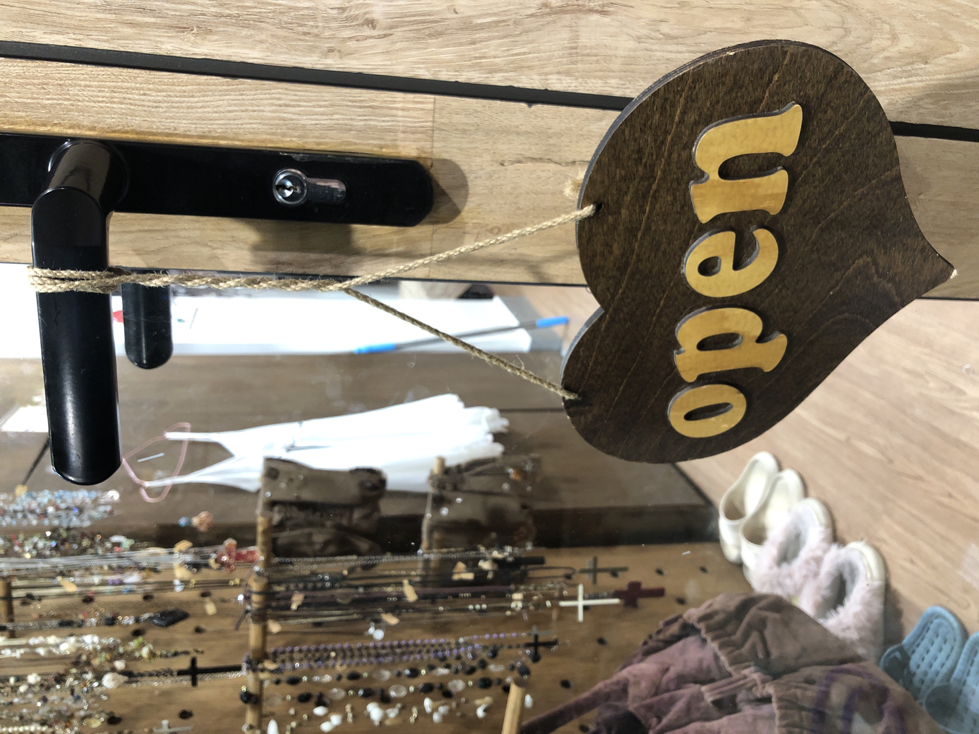 A heart-shaped wooden sign with the word "open" in gold lettering hangs from a door handle, indicating a shop is open. The sign is made of dark wood, with the letters raised and painted a bright gold. The door is made of light-colored wood and has a black metal handle and lock. In the background, a reflection in a mirror shows a white dress hanging on a hanger and various necklaces and jewelry displayed on a wooden stand. Several pairs of slippers are visible on the floor. The image appears to be taken in a shop located in Đà Nẵng, Vietnam.