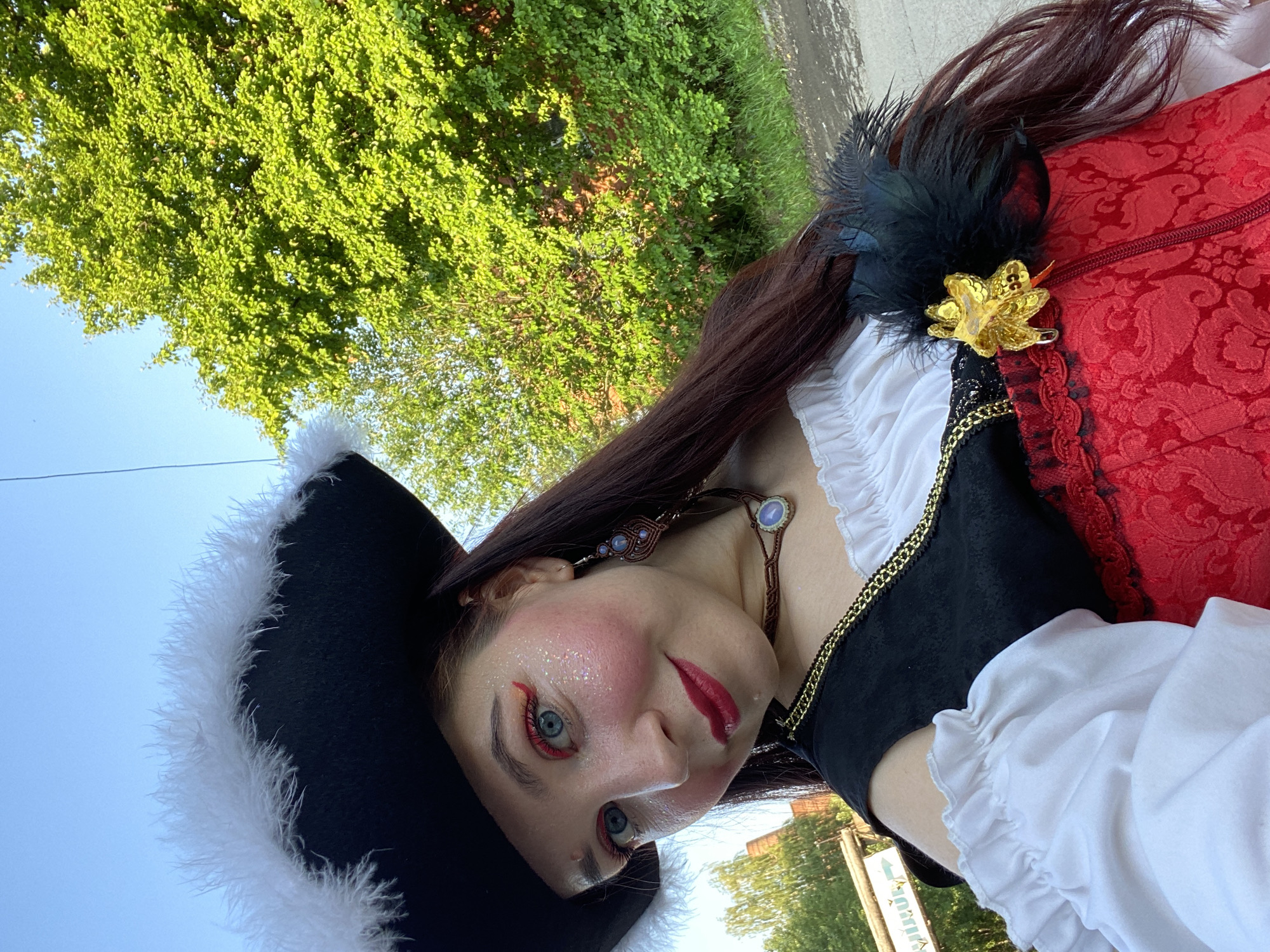 A selfie of a woman dressed as a pirate, wearing a black tricorn hat with white feathers, a white ruffled blouse, a black vest with gold trim, and a red brocade corset with black lace accents. She has blue eyes, red lipstick, and glittery makeup. A gold floral brooch with black feathers is pinned to her shoulder. In the background, there are green trees and a bright blue sky. The location is indicated as Chaikovsky, Russia, with a sign in the background that appears to have Russian lettering and an arrow pointing left. The woman is outdoors, with sunlight illuminating her face and costume.