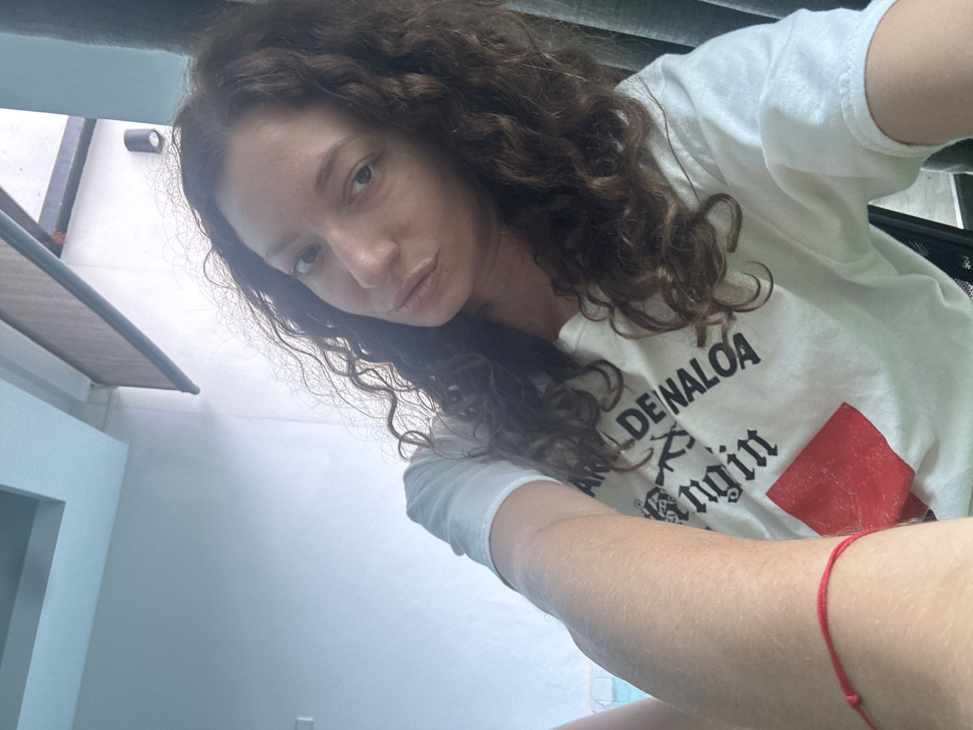 A woman with curly brown hair takes a selfie indoors, with her face angled towards the camera and her left arm extended. She is wearing a white t-shirt with black text and a red square design. A thin red bracelet is visible on her left wrist. The background appears to be a room with light-colored walls and a ceiling that suggests an outdoor or open-air setting, possibly a balcony or patio. The overall lighting is bright, indicating daytime. The location is Ko Samui District, Thailand.