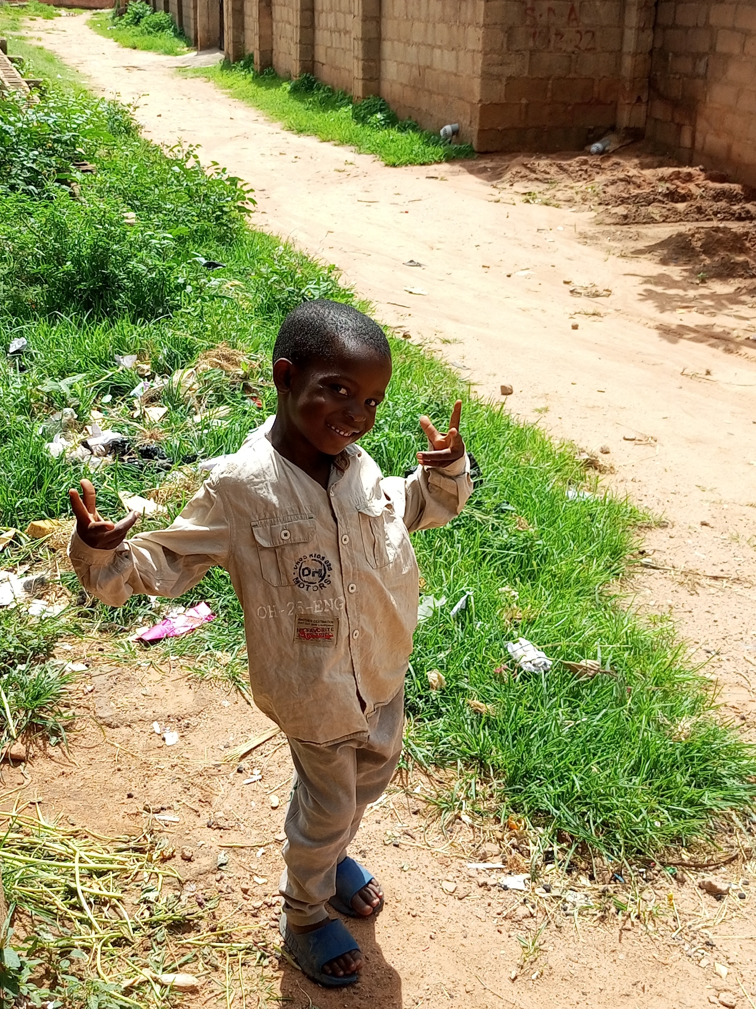 A young boy, appearing to be of Nigerian descent, stands outdoors in what seems to be a dirt road environment in Tempure, Nigeria. He is facing the camera and smiling, holding up two fingers on his right hand in a peace sign gesture. He is wearing a light-colored long-sleeved shirt with pockets and gray pants, and blue sandals. The background consists of a dirt path, green grass, and a brick wall. The overall scene suggests a casual, everyday moment in a developing urban or semi-urban area.