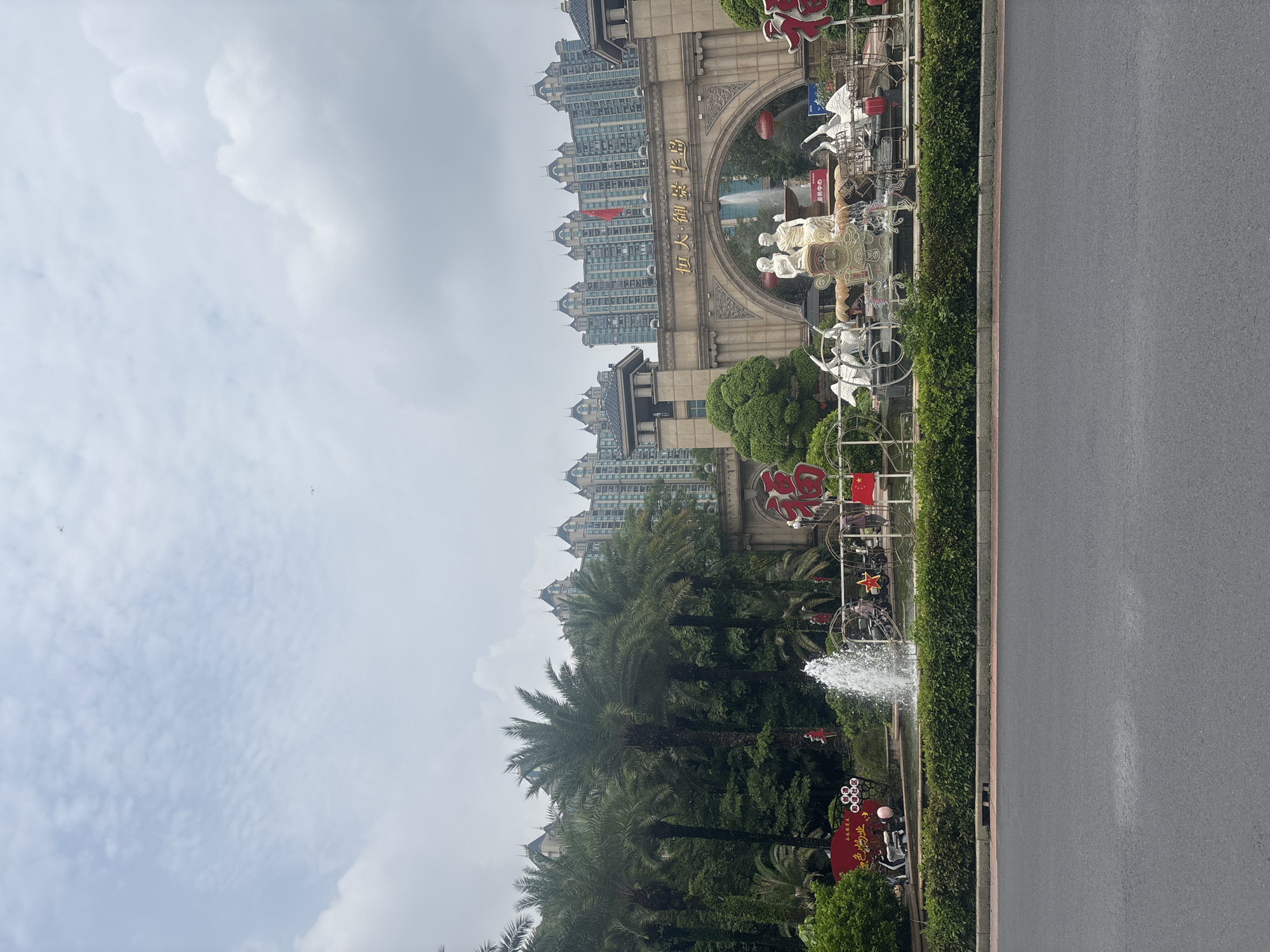 The image depicts a street view in Nanhai District, China, featuring a grand entrance archway adorned with Chinese characters. Tall apartment buildings with ornate roofs form the backdrop. In the foreground, a fountain sprays water, and various decorative sculptures, including human figures and swans, surround it. Red lanterns and banners, some with Chinese characters, are visible, suggesting a festive occasion. Palm trees and lush green bushes line the area. A road and sidewalk are in the lower portion of the image. Two small airplanes are faintly visible in the overcast sky.