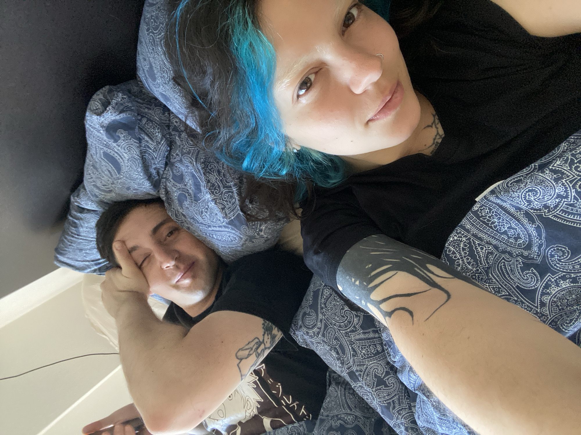 A selfie shows two people lying in bed under a blue and white patterned duvet and pillows. The person in the foreground has dark hair with streaks of blue, a nose ring, fair skin, and is wearing a black shirt. A tattoo is visible on their neck, and a large dark tattoo is on their forearm. The other person in the background is fair-skinned, with dark hair, and wearing a black shirt. They are resting their head on their hand. A tattoo is visible on their upper arm. The location cannot be determined from the image alone.